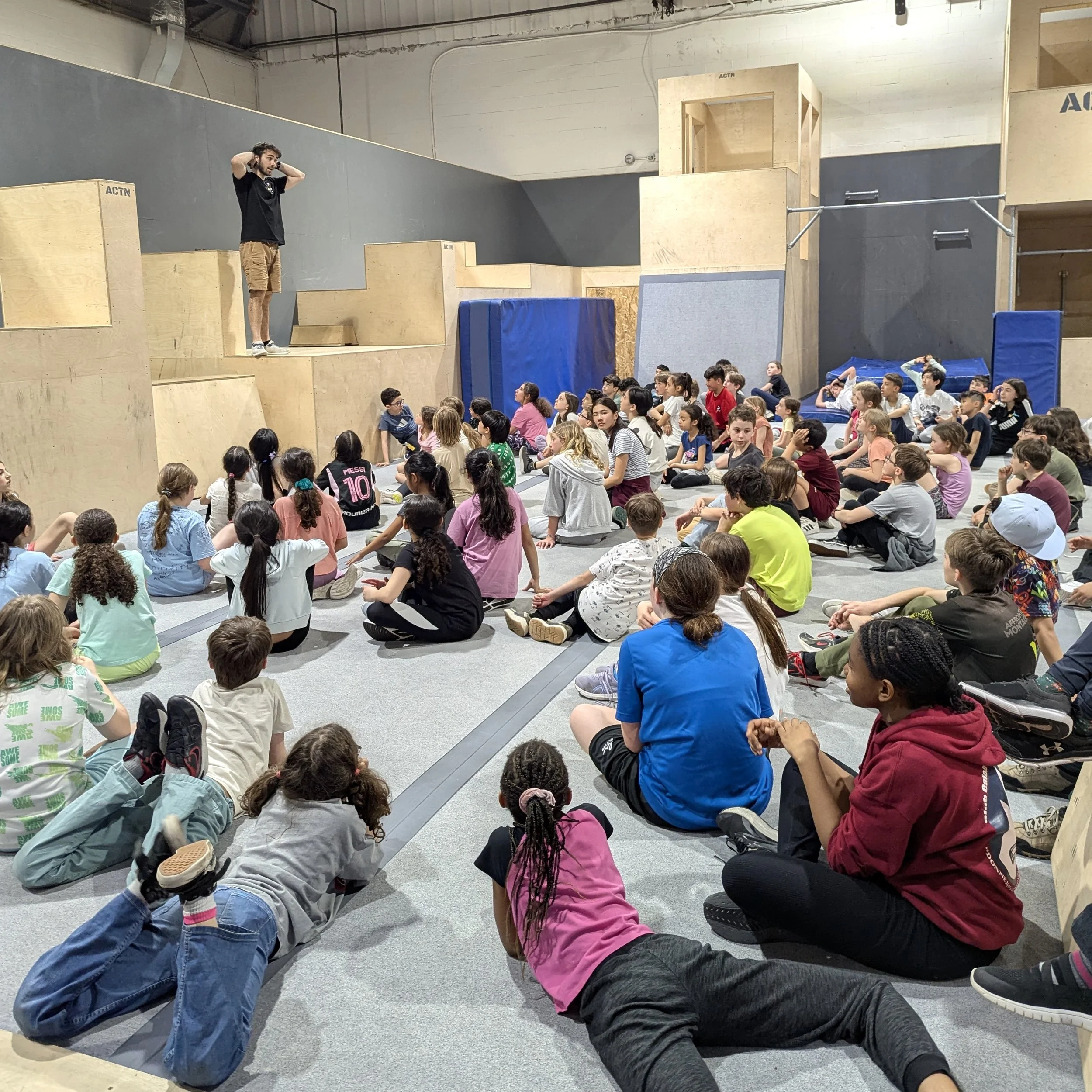 Un groupe d'enfants assis sur le sol d'une salle d'escalade intérieure, regardant un instructeur debout sur une plateforme en bois.