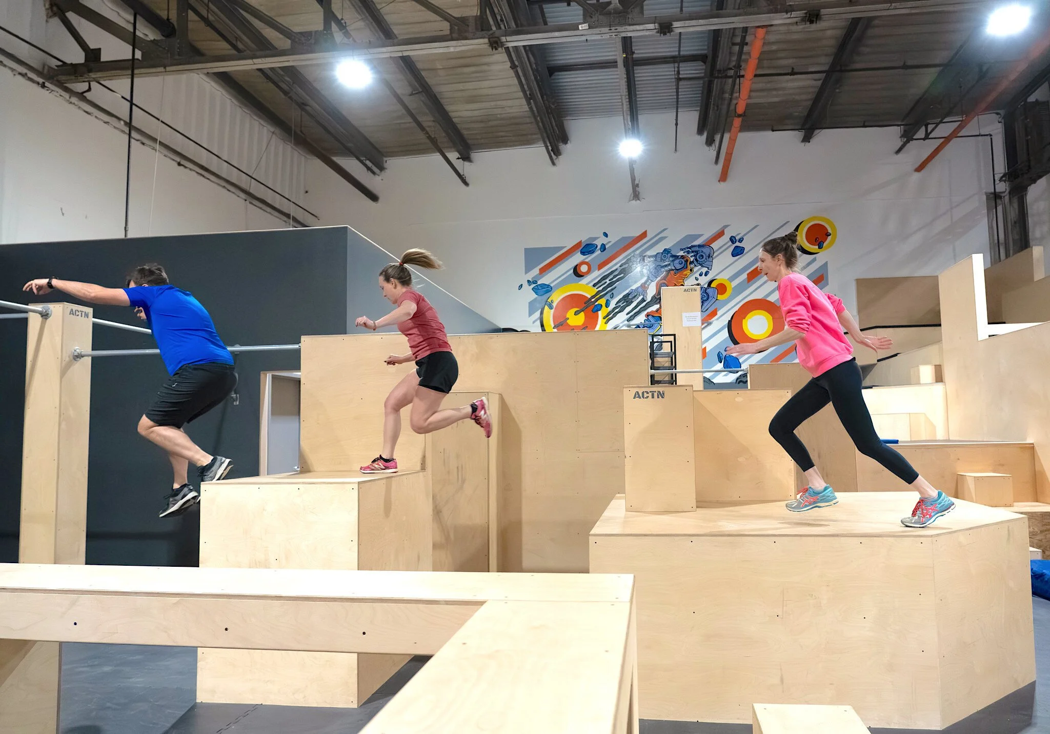Trois personnes pratiquent le parkour à l'intérieur sur des plateformes en bois, dans une salle avec un plafond industriel et un mur décoré d'une fresque colorée.