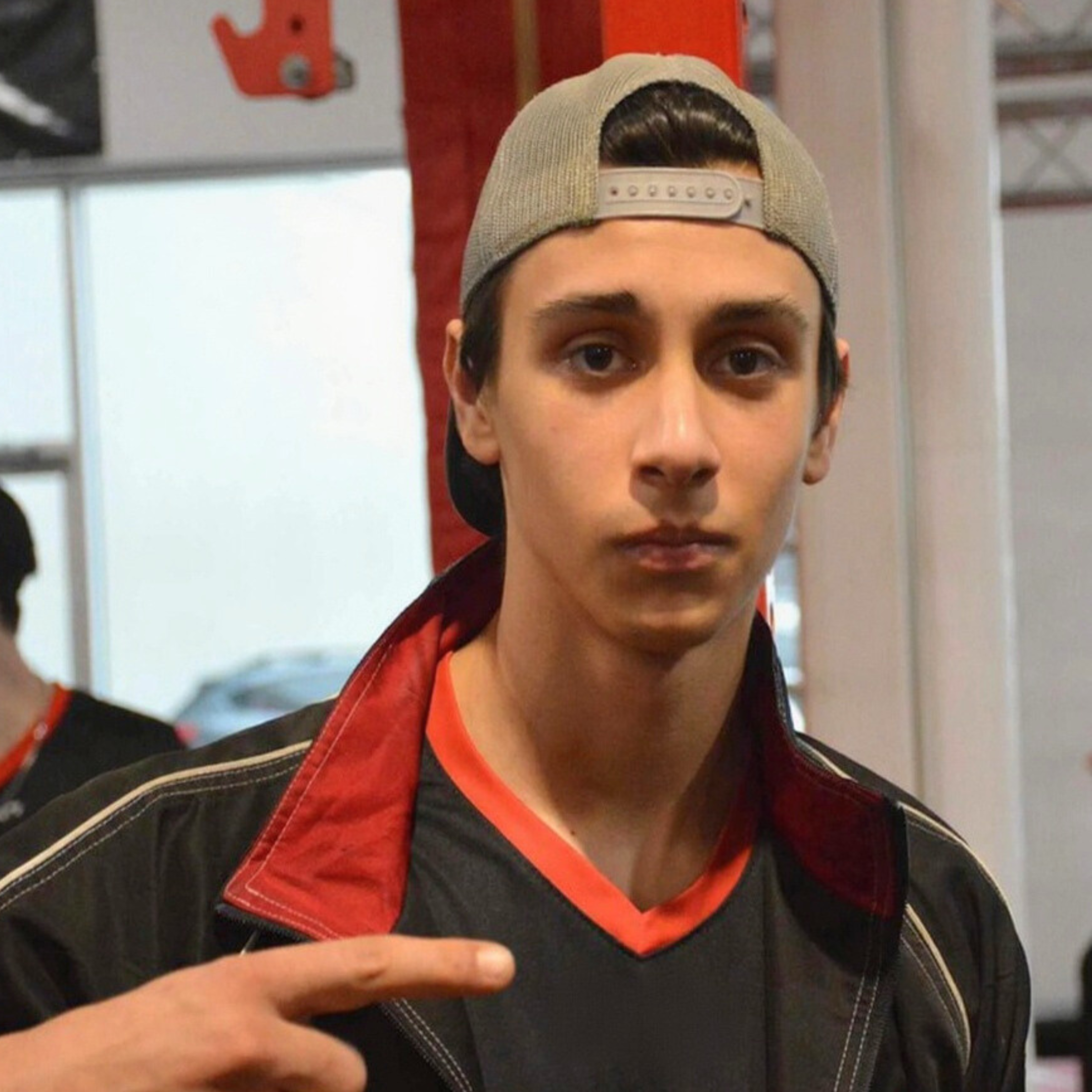 Jeune homme portant une casquette à l'envers, veste noire avec détails rouges, pointant vers lui-même avec son doigt