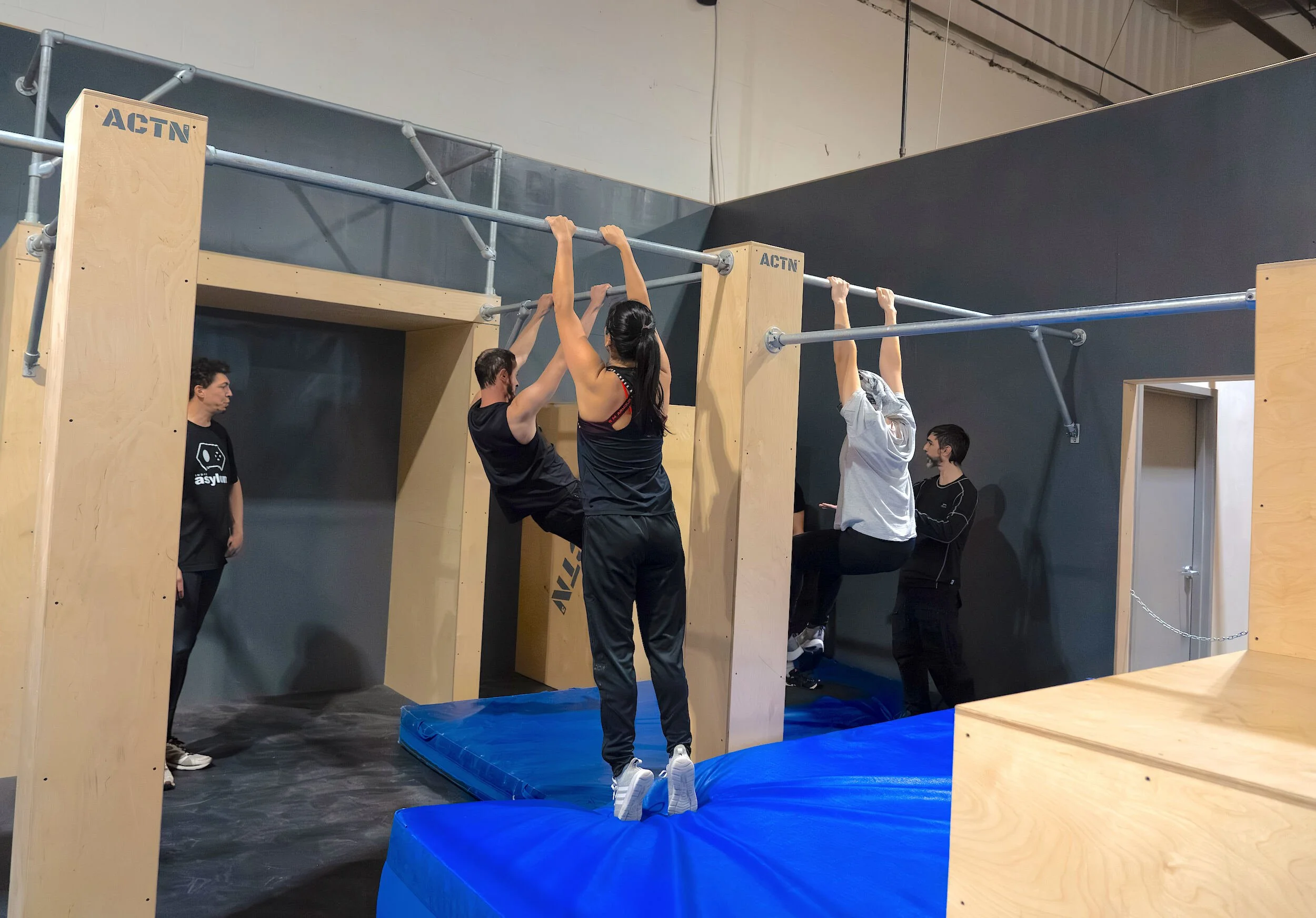 Groupe de jeunes personnes faisant de la musculation sur une barre fixe dans une salle d'entraînement, avec un homme en train d'observer.
