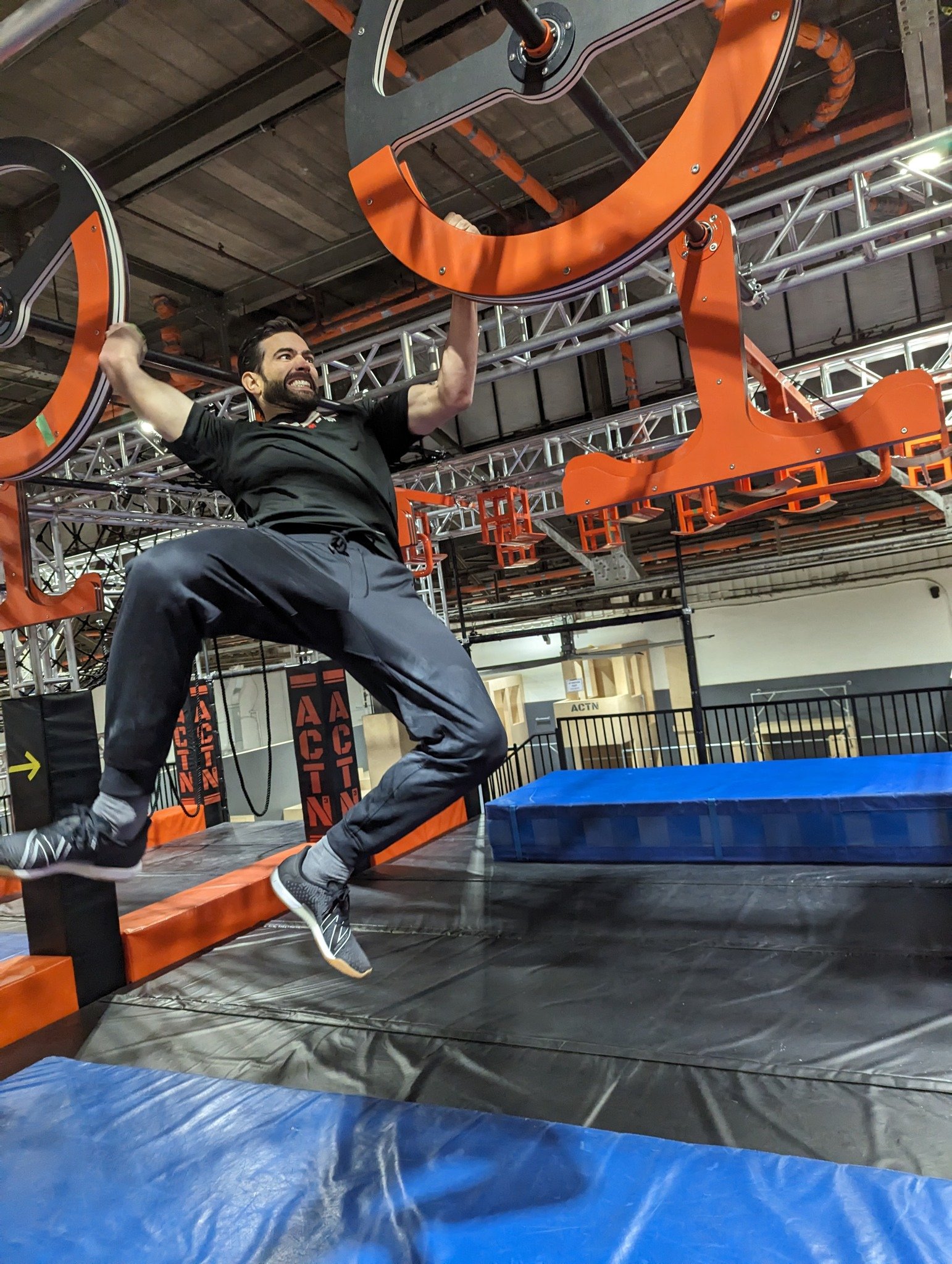 Un homme souriant et énergique en train de franchir une obstacle dans une structure de Ninja Warrior sur deux étages, équipé de structures métalliques et de tapis de sécurité.