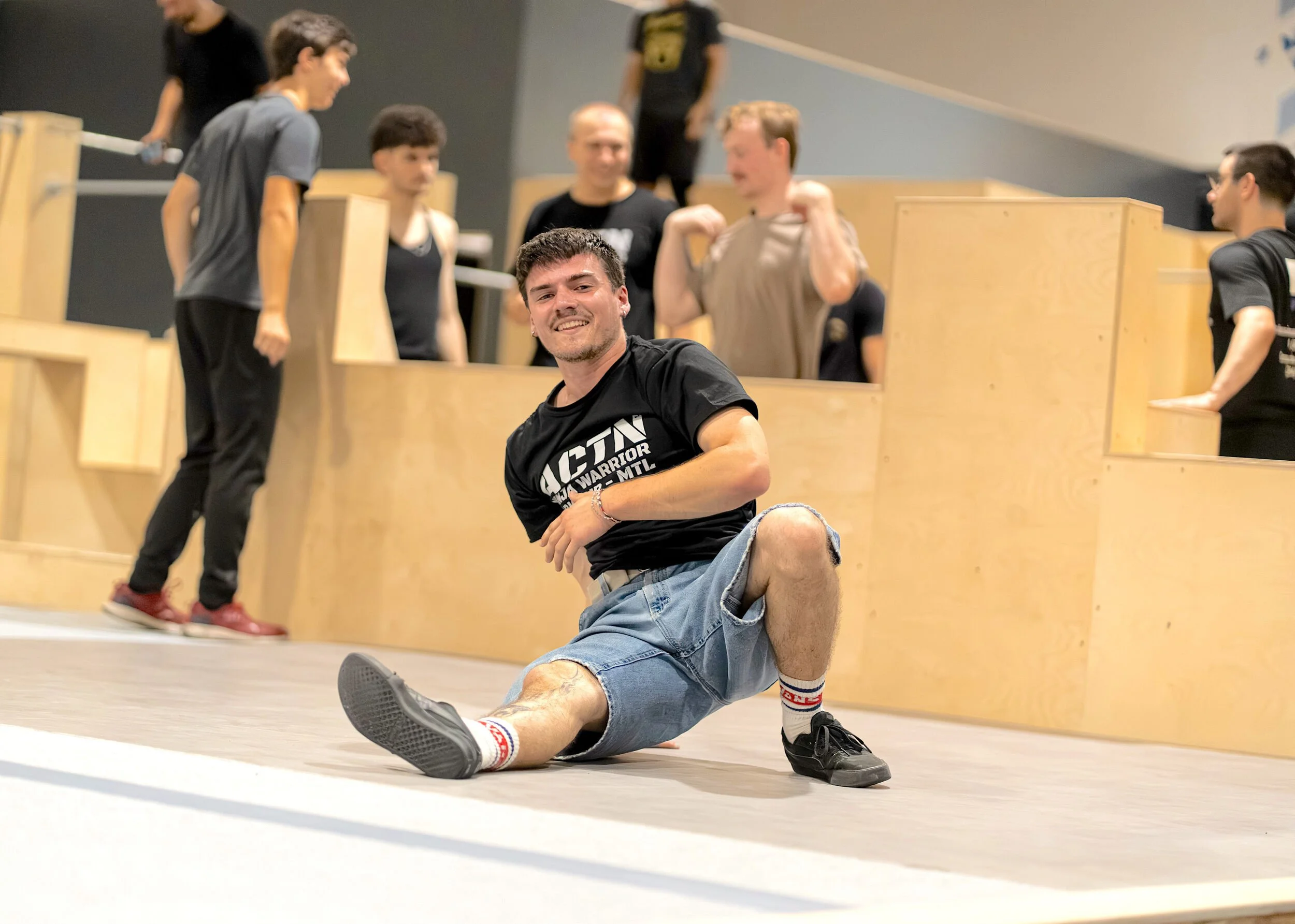 Un jeune homme souriant fait des étirements en position assise par terre dans une salle de sport avec plusieurs autres jeunes en arrière-plan.