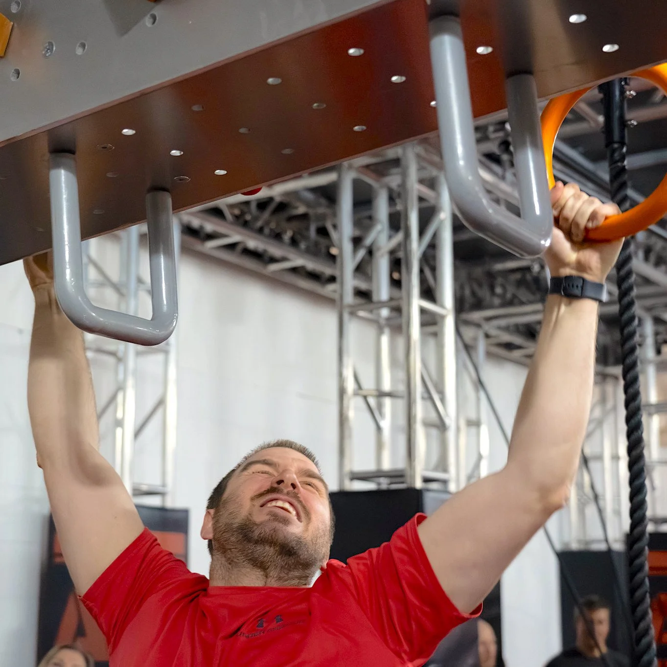 Un homme en t-shirt rouge s'accroche à des poignées métalliques lors d'une compétition de type parcours d'obstacles, avec un visage montrant de l'effort.