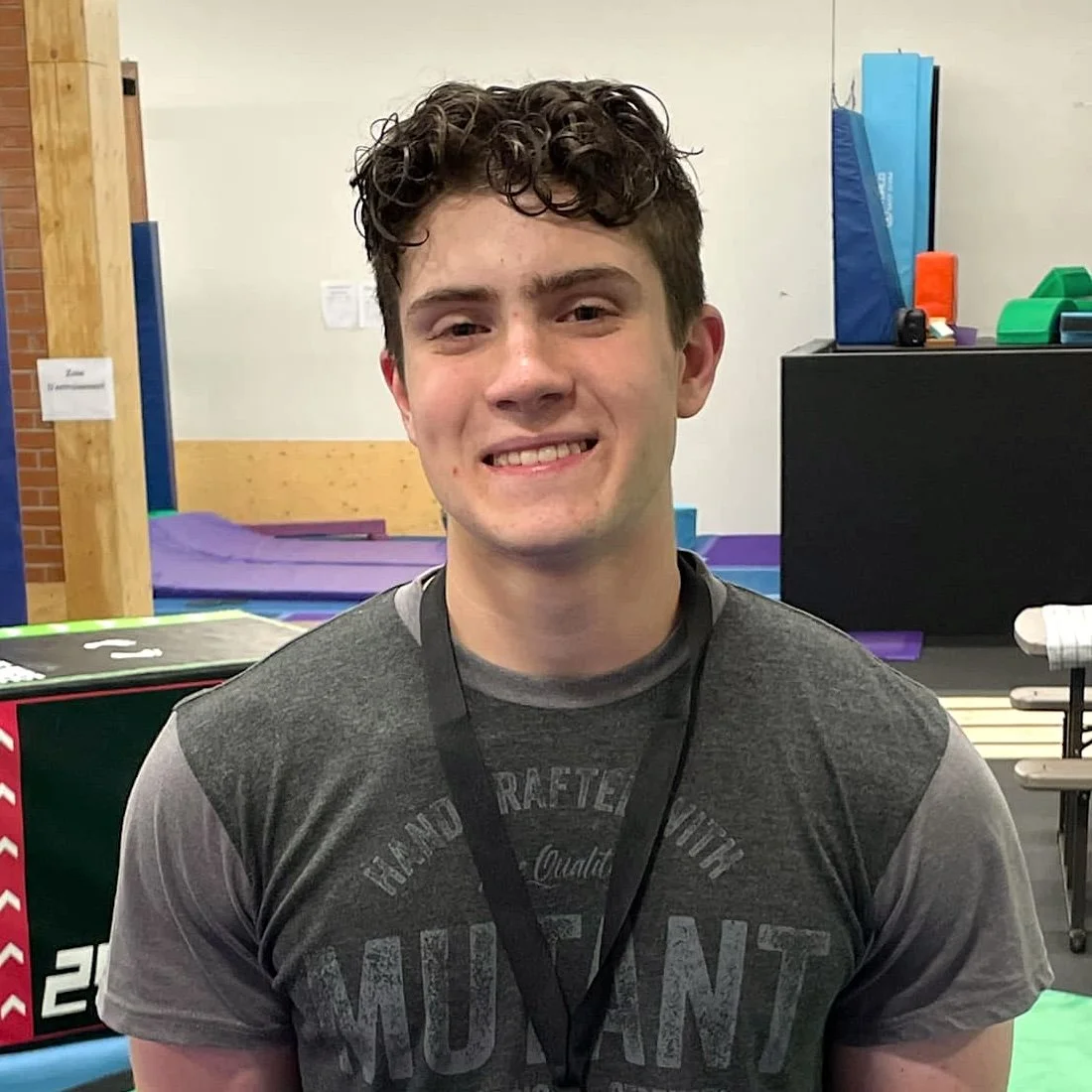 Jeune homme souriant avec des cheveux bouclés, portant un t-shirt gris foncé, dans une salle de sport ou une salle de loisirs équipée de tapis de gymnastique et de protections.