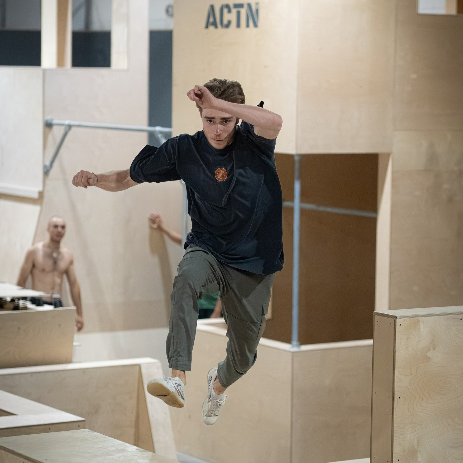 Un jeune homme court dans une salle d'entraînement intérieur en bois, avec un mur portant le logo ACTN3, en portant un t-shirt noir et un pantalon marron, faisant du parkour ou de la course d'obstacles.