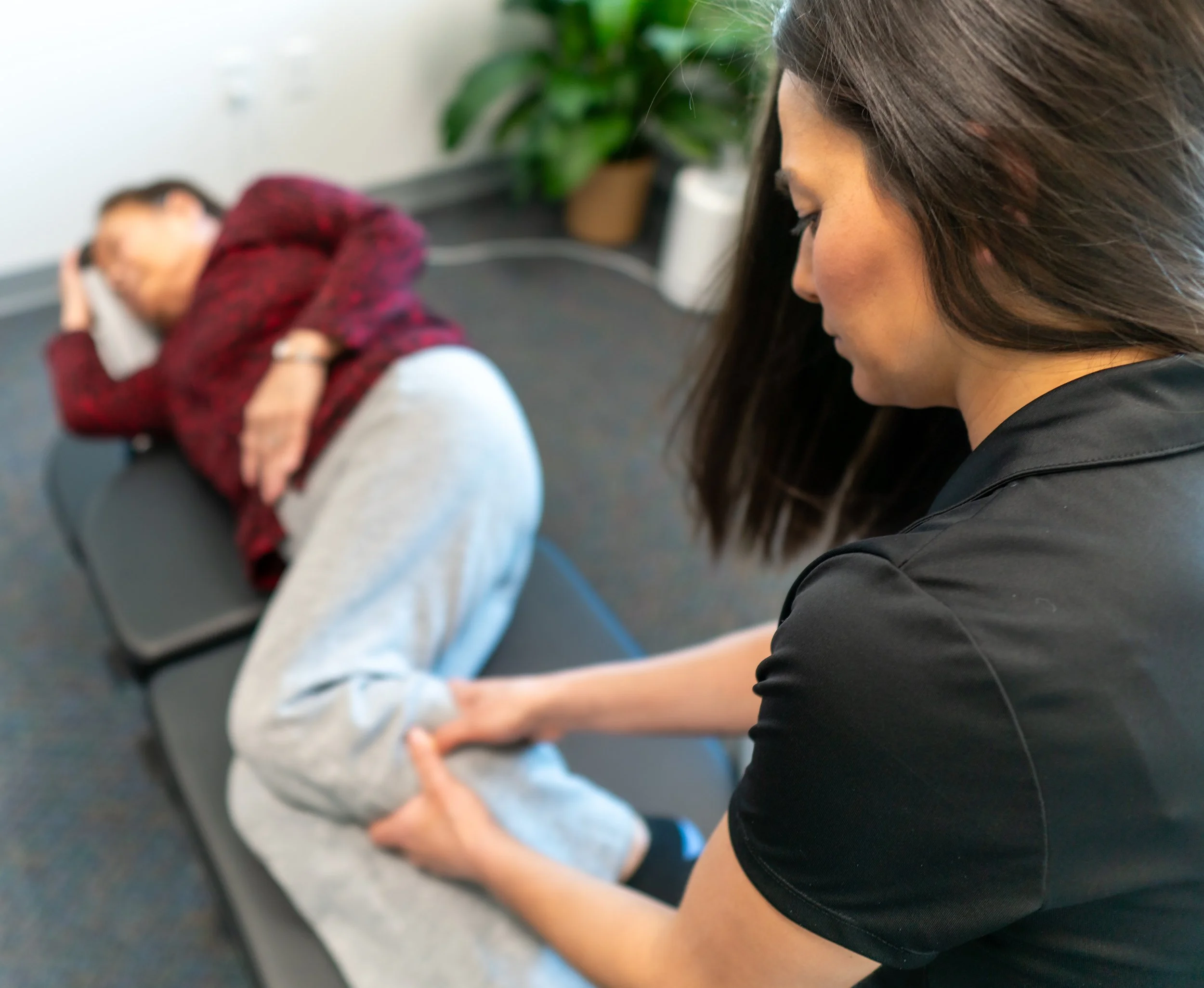 A woman with long dark hair, wearing a black shirt, is smiling and providing chiropractic care to an elderly woman lying on a massage table. The elderly woman is wearing a blue shirt and has light-colored hair. A world map decoration is visible on the wall in the background.