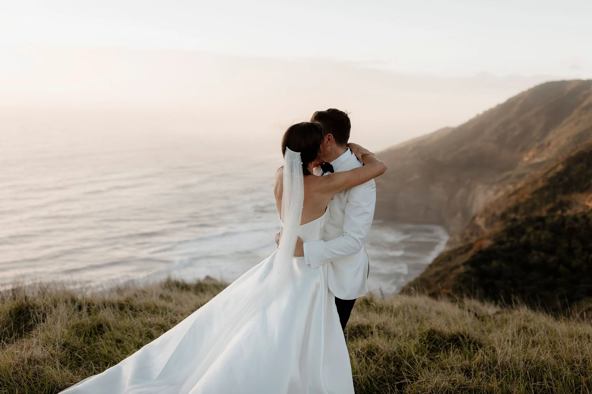 A bride abd groom embracing on the edge of a cliff with a beautiful view of the coastline below them 