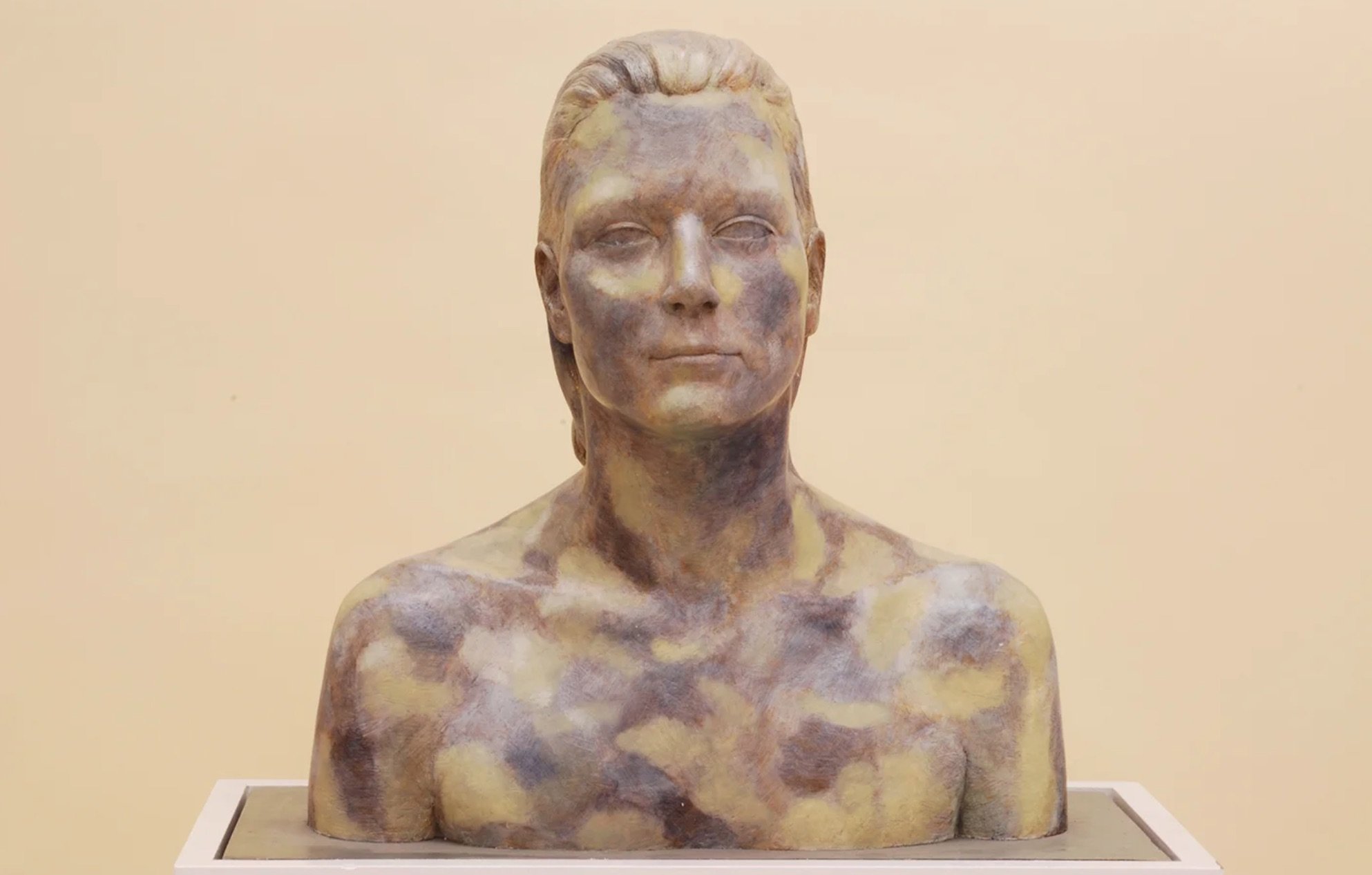 A bust sculpture of a woman with closed lips, high cheekbones, and styled hair, painted in an abstract, multicolored pattern with earthy tones.