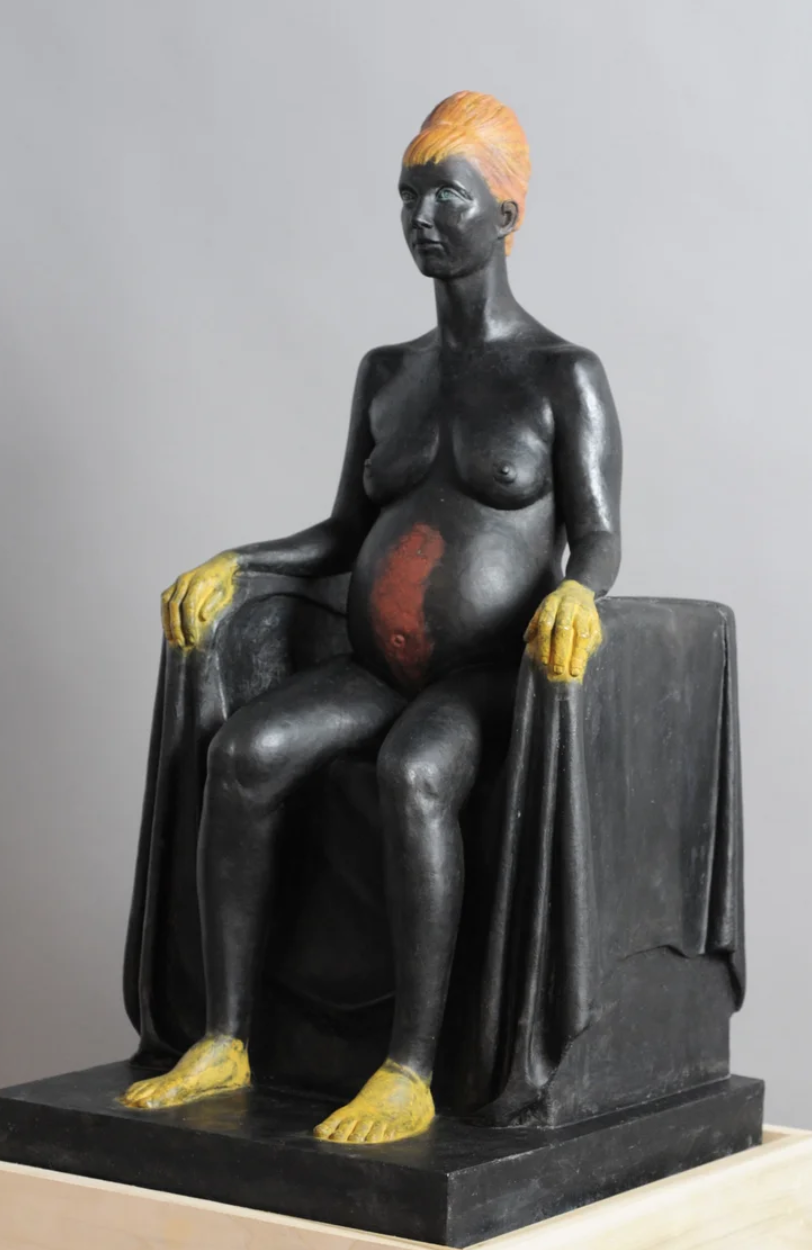 Sculpture of a pregnant woman with painted yellow hair, yellow hands and feet, seated on a dark stool, with a blackened body and an orange-red area on her abdomen, against a plain gray background.