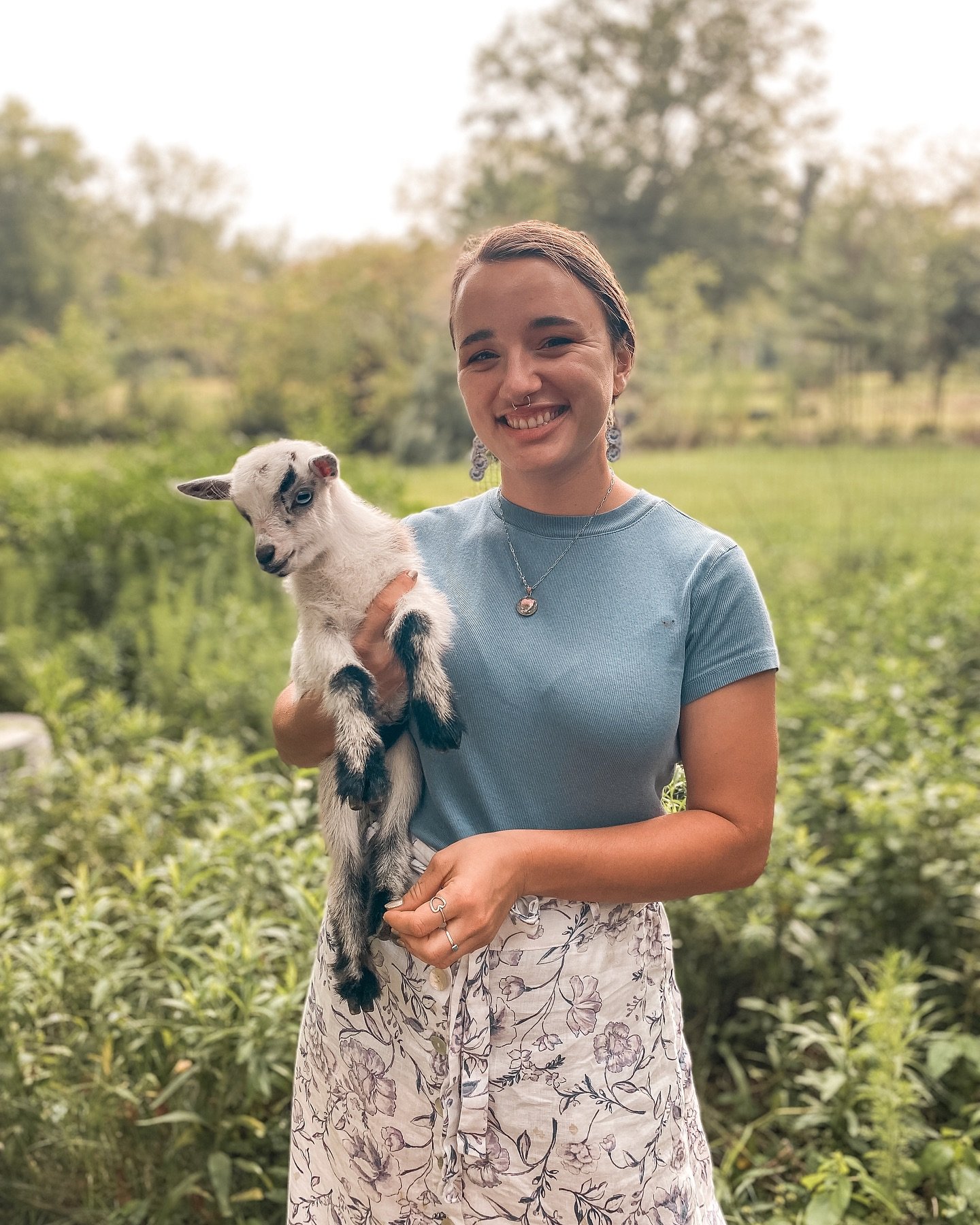 A young woman with short hair, wearing a light blue t-shirt, floral skirt, and jewelry, holding a small white goat with blue eyes in a lush green outdoor setting during daytime.