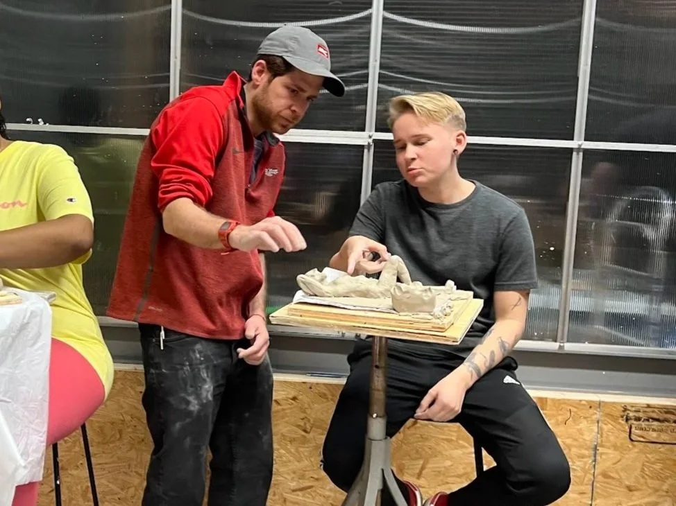 Two men are engaged in a pottery lesson or class, working with clay sculptures, in a room with metal-framed windows.
