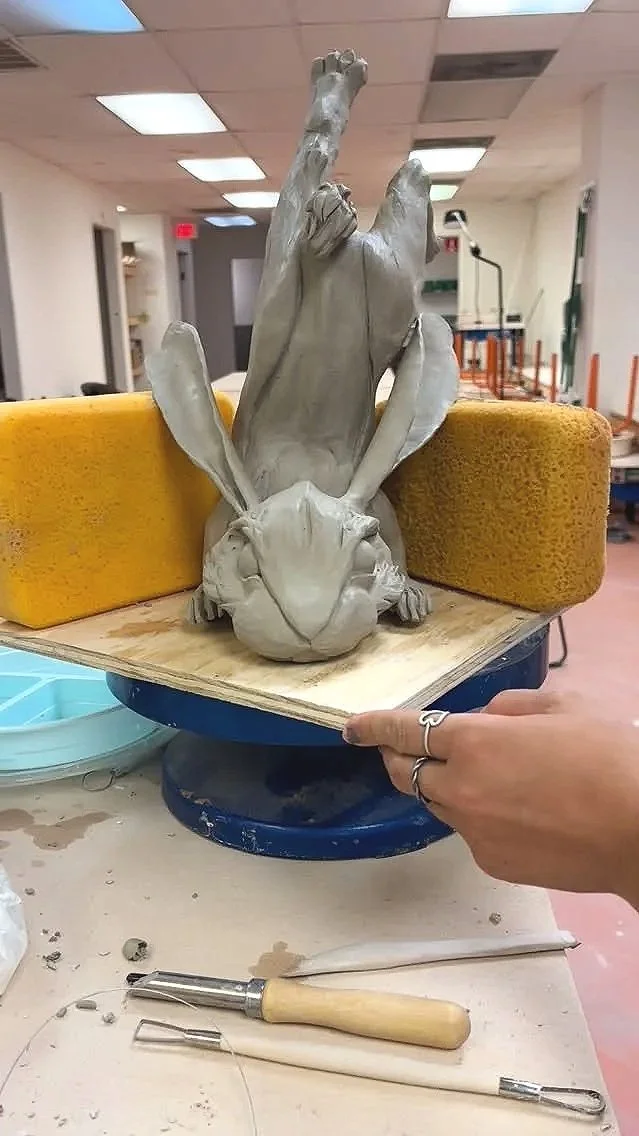 A sculpture of a rabbit lying on its back with its legs up, in progress, on a turntable, in an art studio.