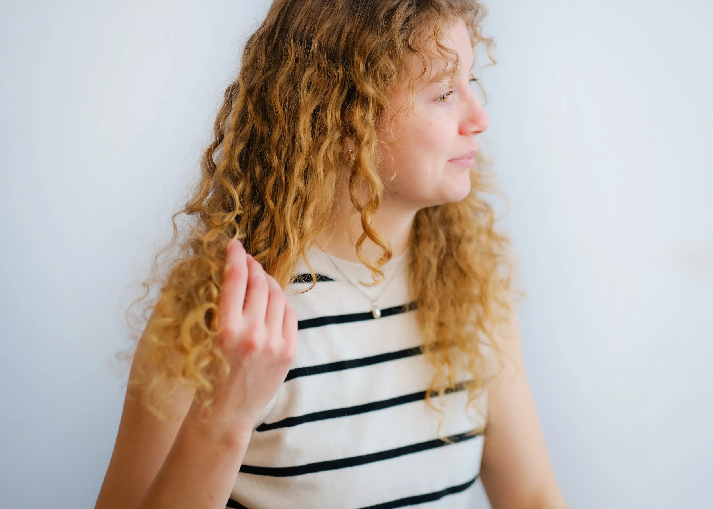 A woman with curly red hair wearing a white and black striped shirt, standing sideways and touching her hair while looking to the right.