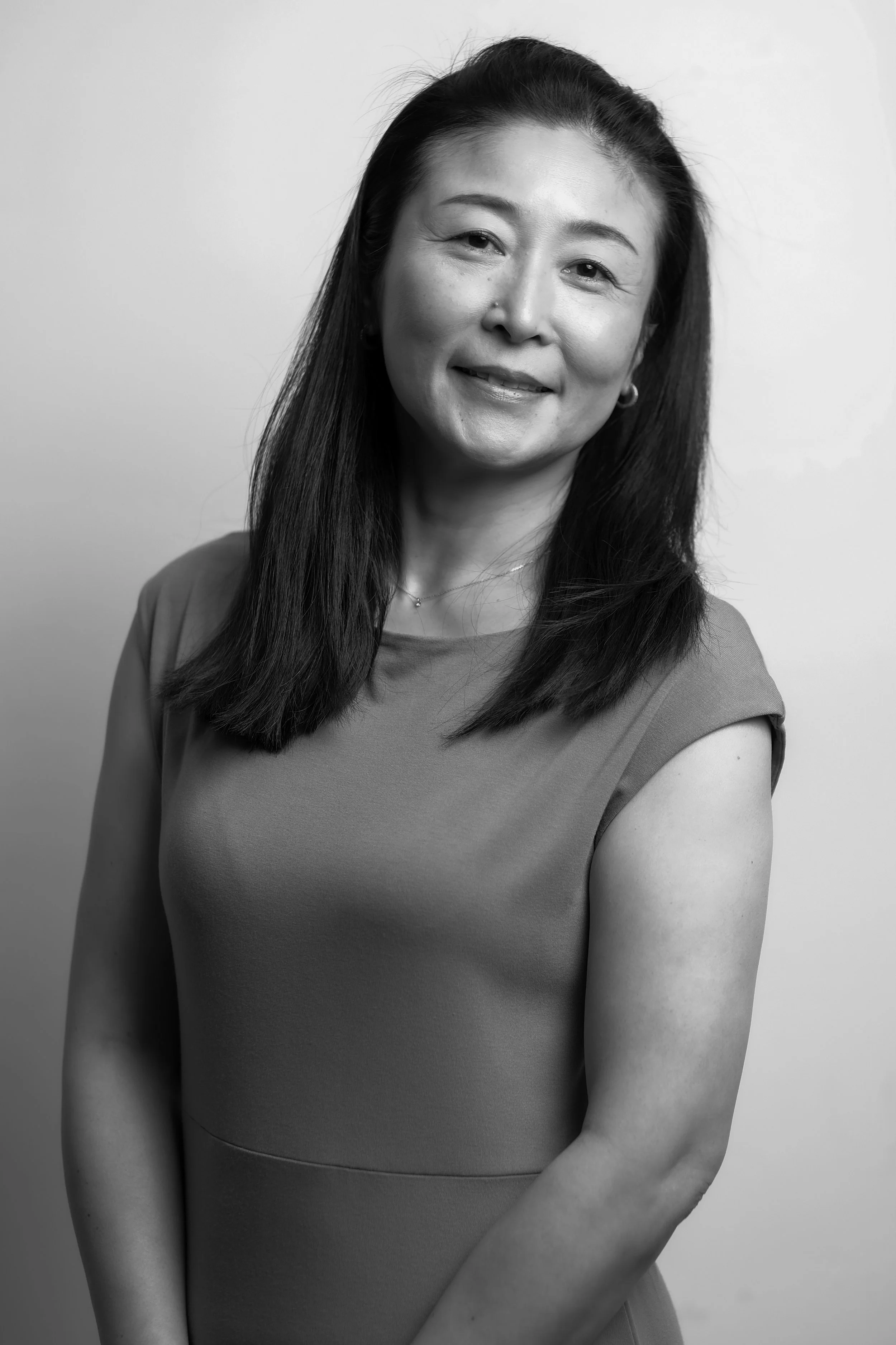 A black-and-white portrait of a woman with long dark hair, smiling slightly, wearing a short-sleeved dress, standing against a plain background.
