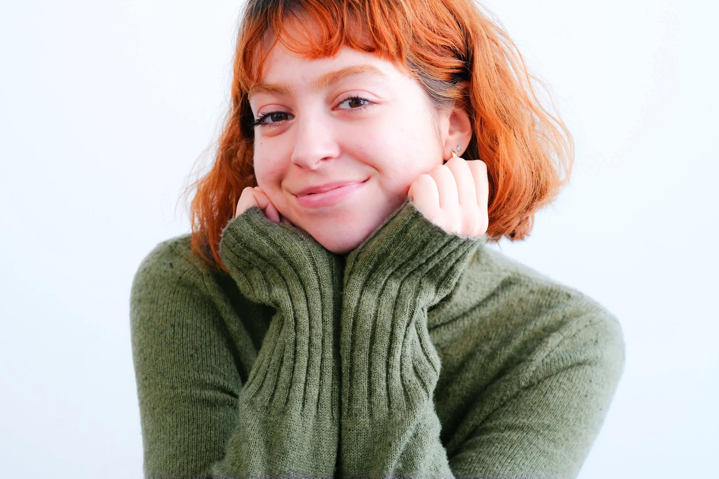 Close-up of a young woman with short, curly red hair, smiling gently, wearing a green sweater, with her hands resting on her cheeks against a plain light background.