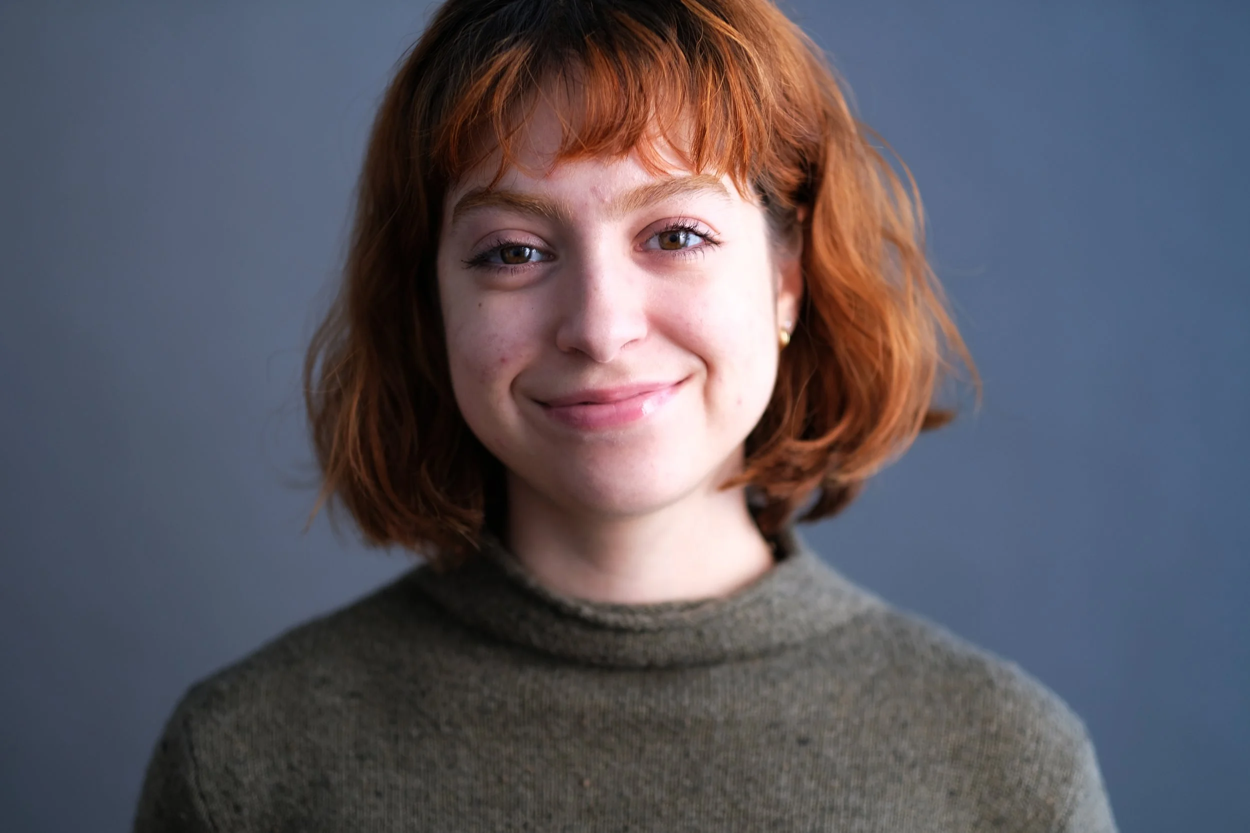 A young woman with shoulder-length red hair, light skin, wearing a dark green turtleneck, smiling against a solid gray background.