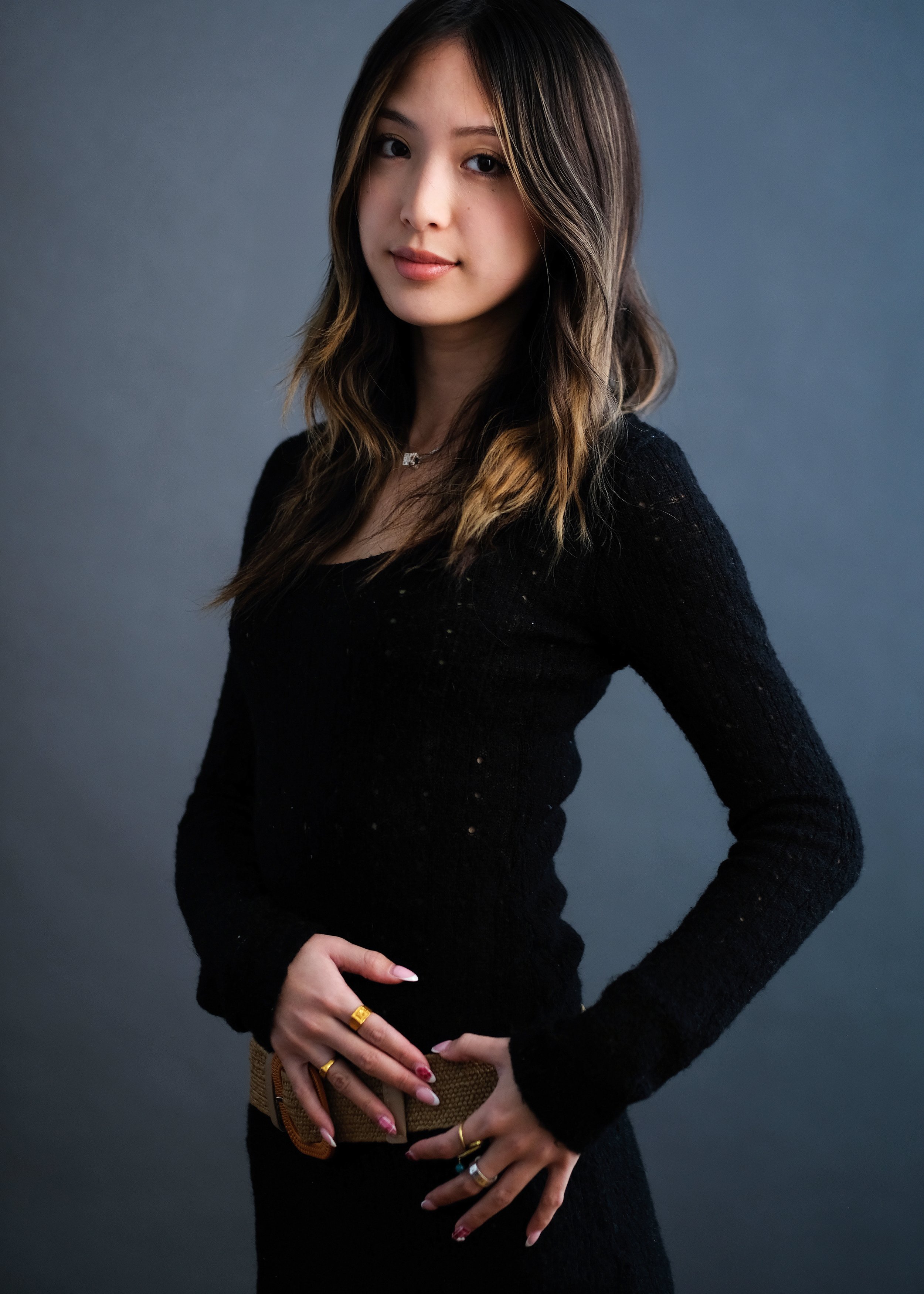 A woman with long wavy brown hair, wearing a black sweater, standing against a gray background.