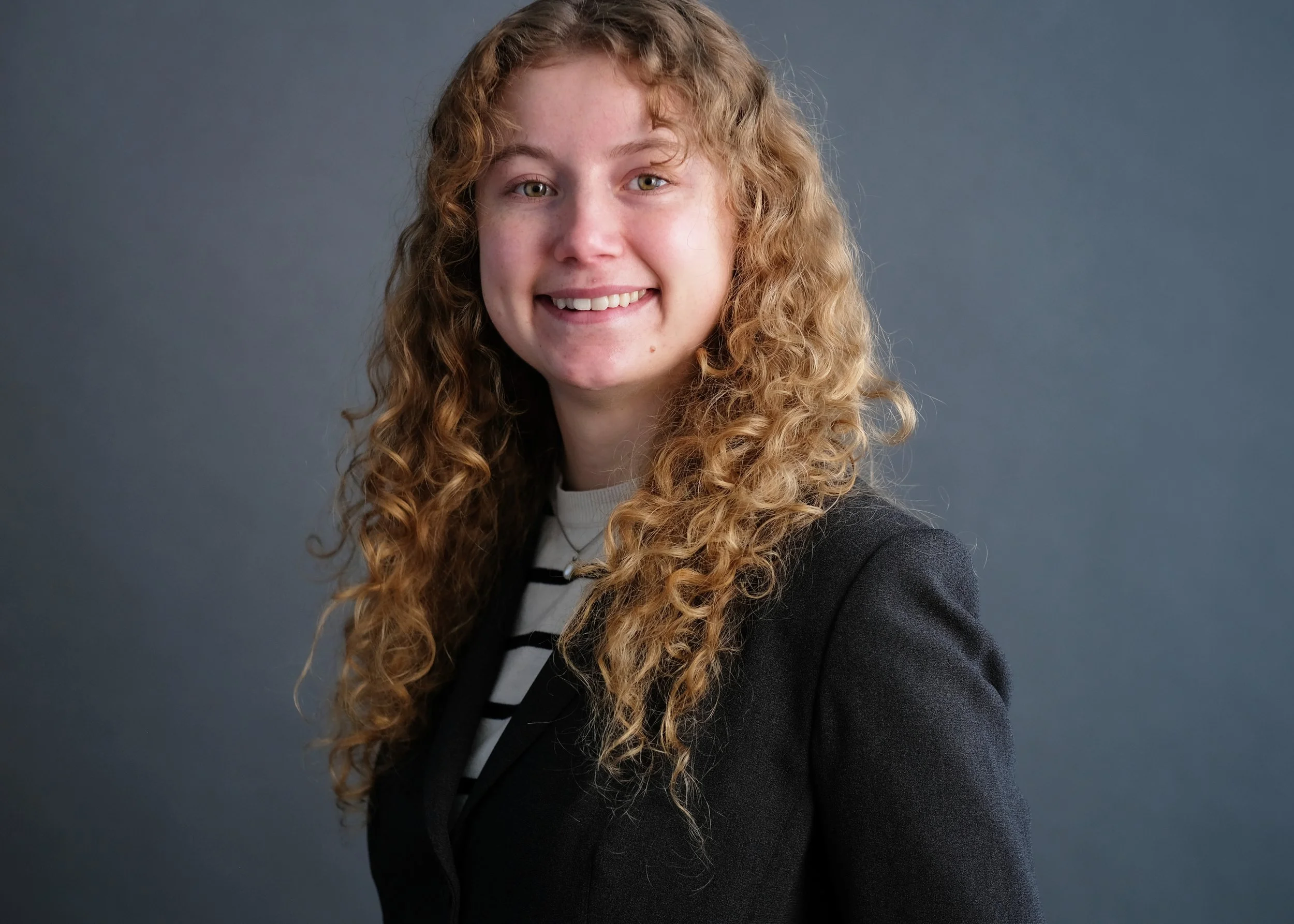 A woman with long, curly red hair, wearing a dark blazer over a white and black striped shirt, smiling at the camera against a gray background.
