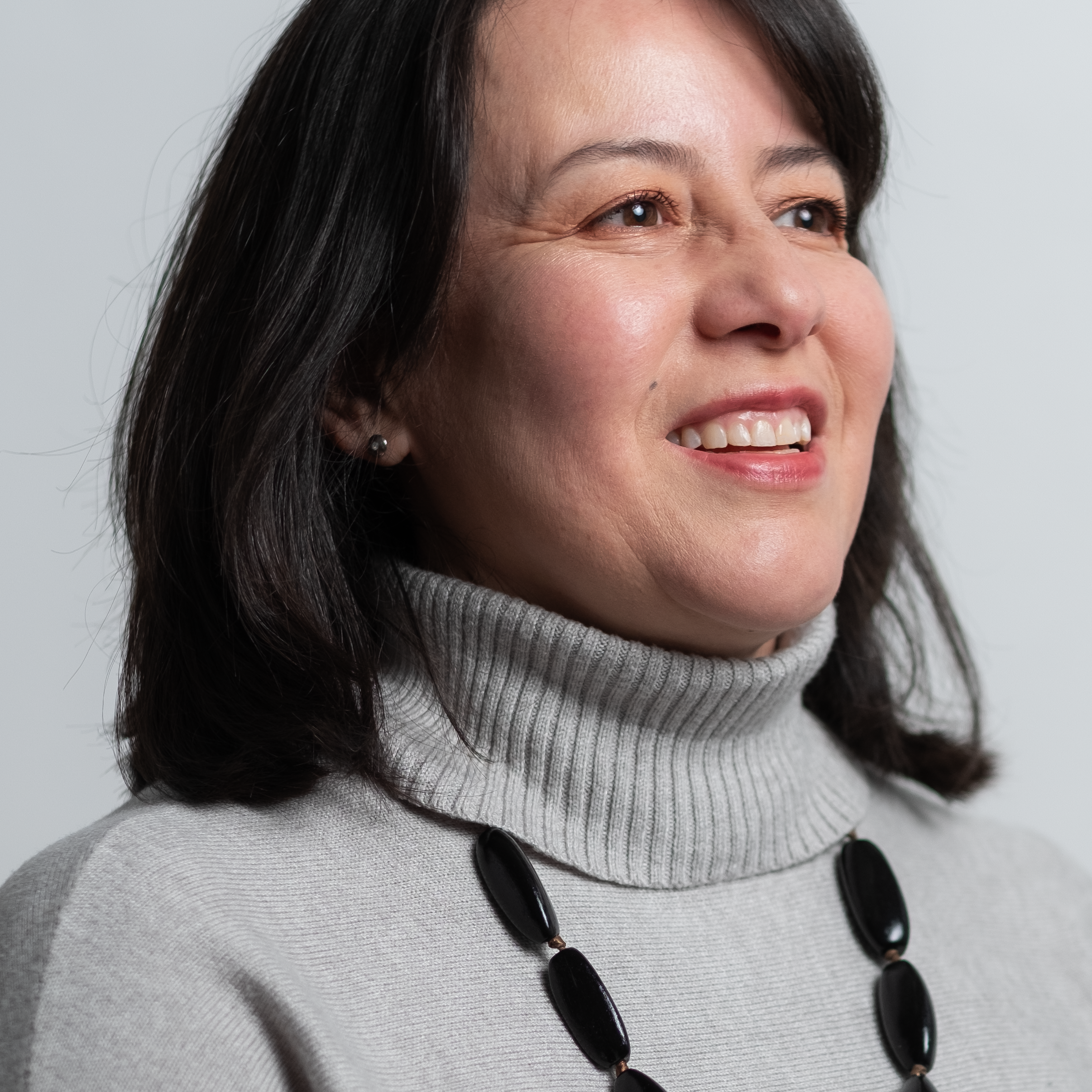 Close-up of a woman smiling with dark hair, wearing a gray turtleneck and a black beaded necklace.