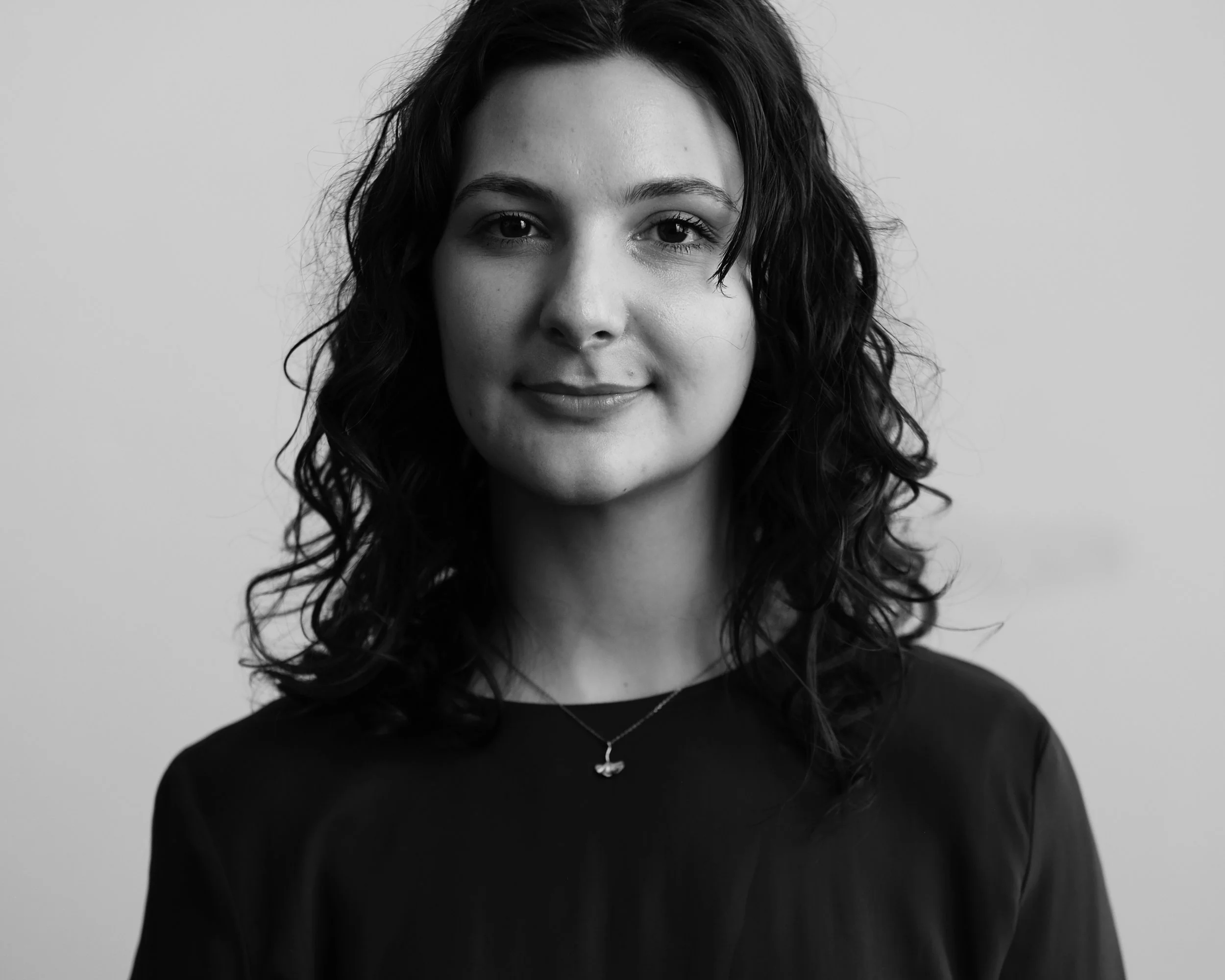 A black and white portrait of a smiling woman with wavy dark hair, wearing a dark top and a small necklace, against a plain light background.
