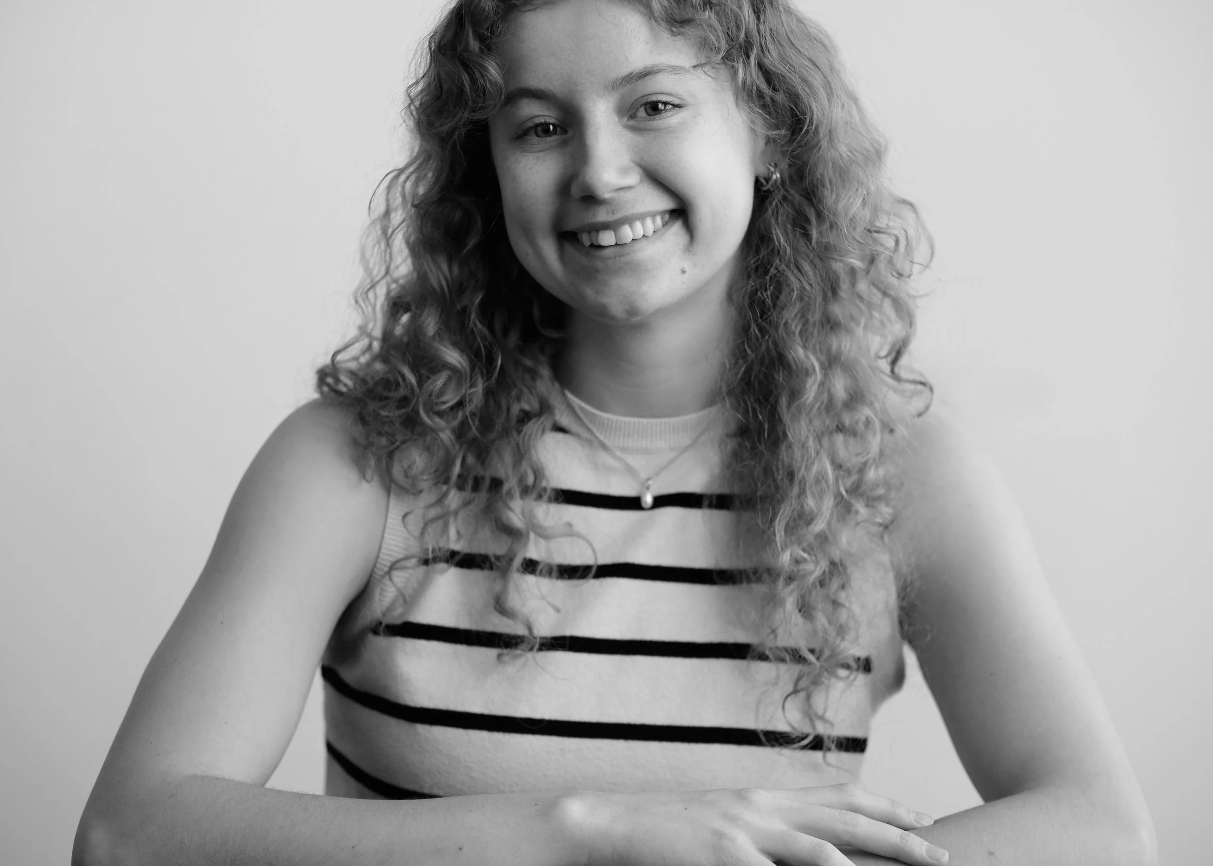 Black and white portrait of a young woman with curly hair smiling, wearing a striped sleeveless top and a necklace, sitting with her hands resting on her lap against a plain background.