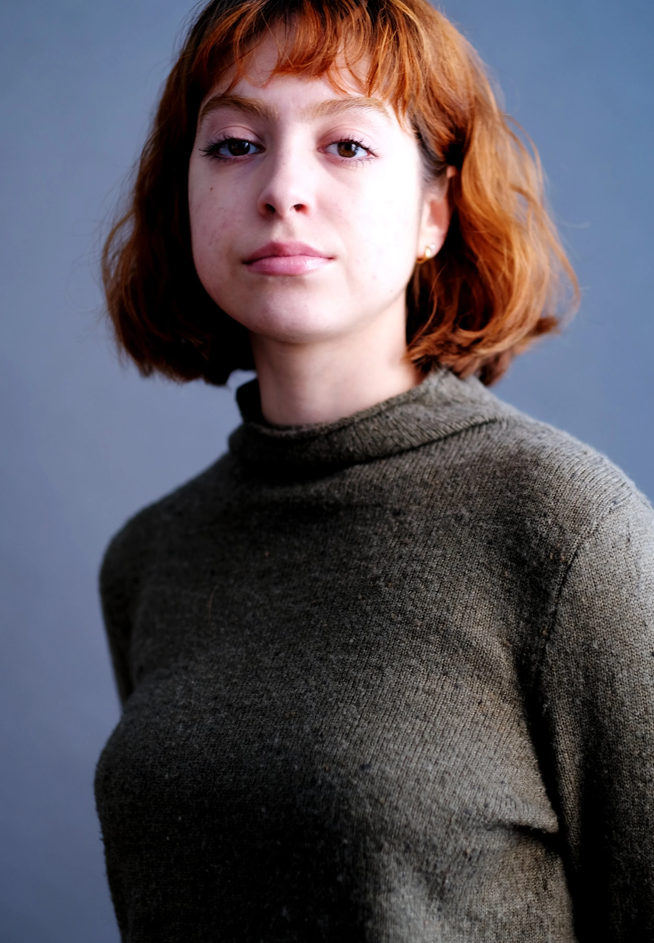 A young woman with short red hair and fair skin, wearing a dark green turtleneck sweater, looking into the camera with a neutral expression, against a blue background.