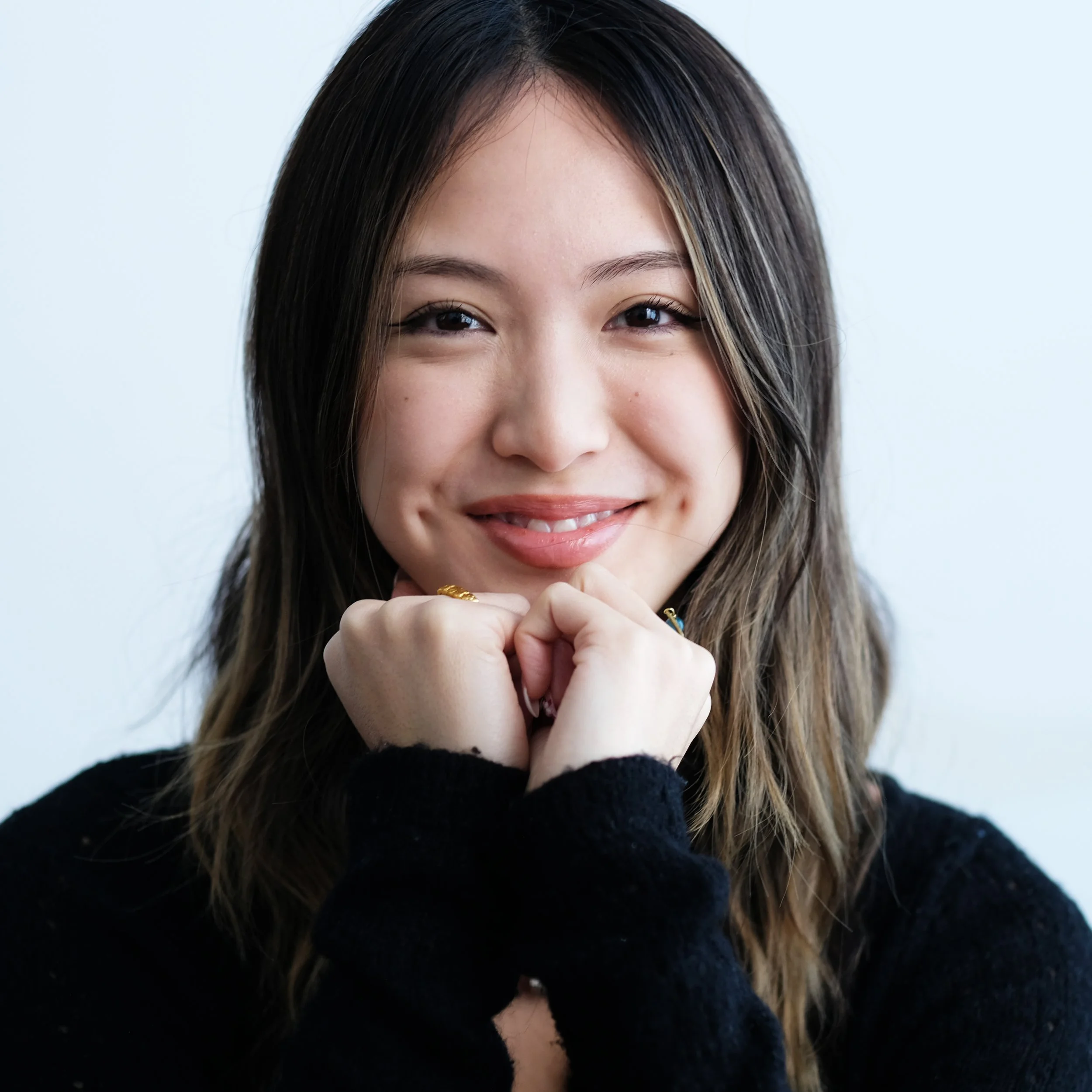 Close-up of a young woman with long brown hair and a warm smile, resting her chin on her hands, wearing a black sweater against a plain light background.