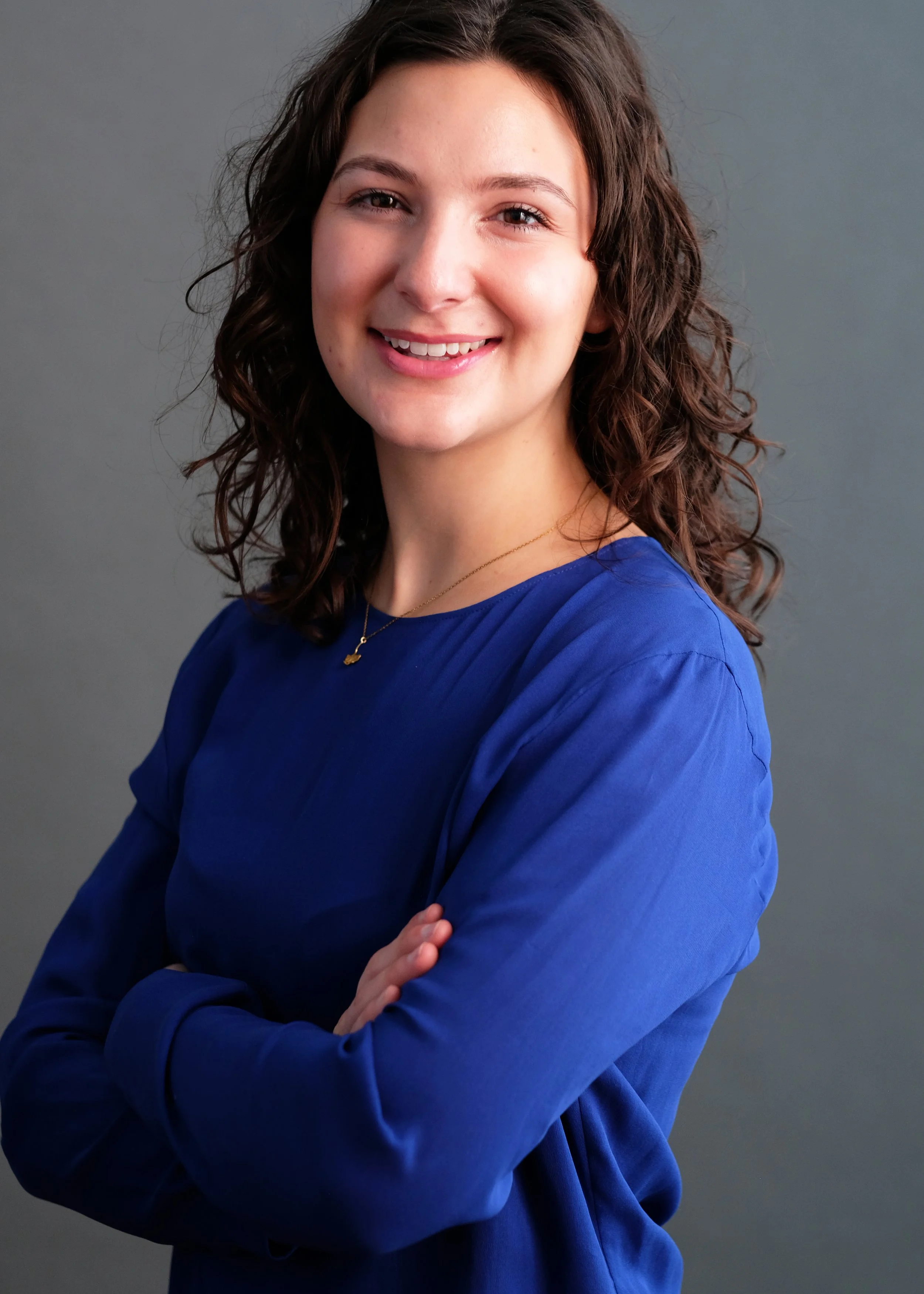 A woman with shoulder-length curly brown hair smiling and wearing a blue long-sleeve top, standing against a gray background.