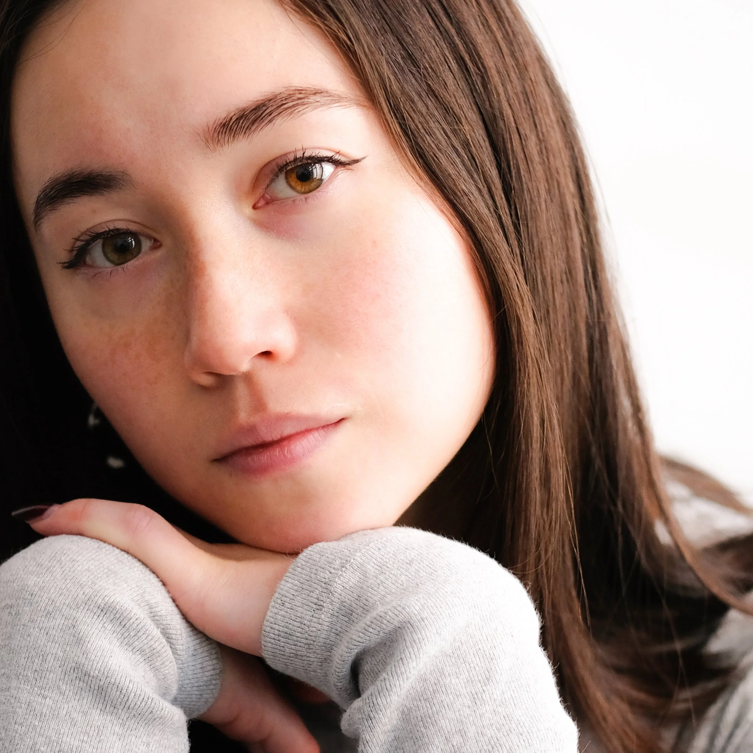 Close-up of a young woman with brown hair and light skin resting her chin on her hand, looking at the camera.