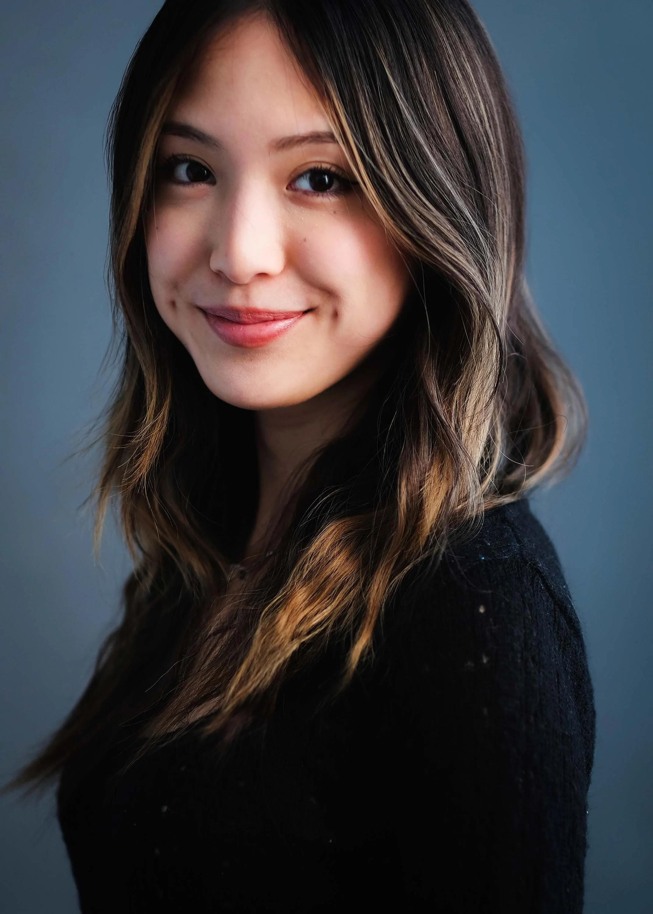 Close-up portrait of a young woman with long, wavy brown hair and a warm smile, wearing a black top, against a blue background.