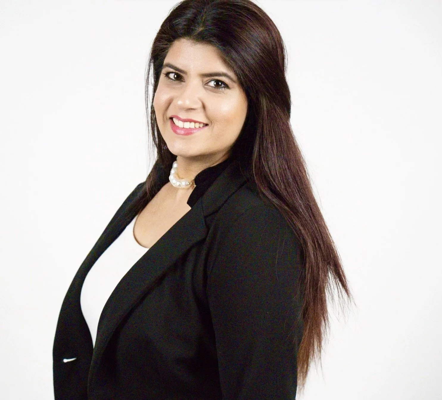 A woman with long brown hair smiling at the camera, wearing a black blazer, white top, and a pearl necklace against a white background.