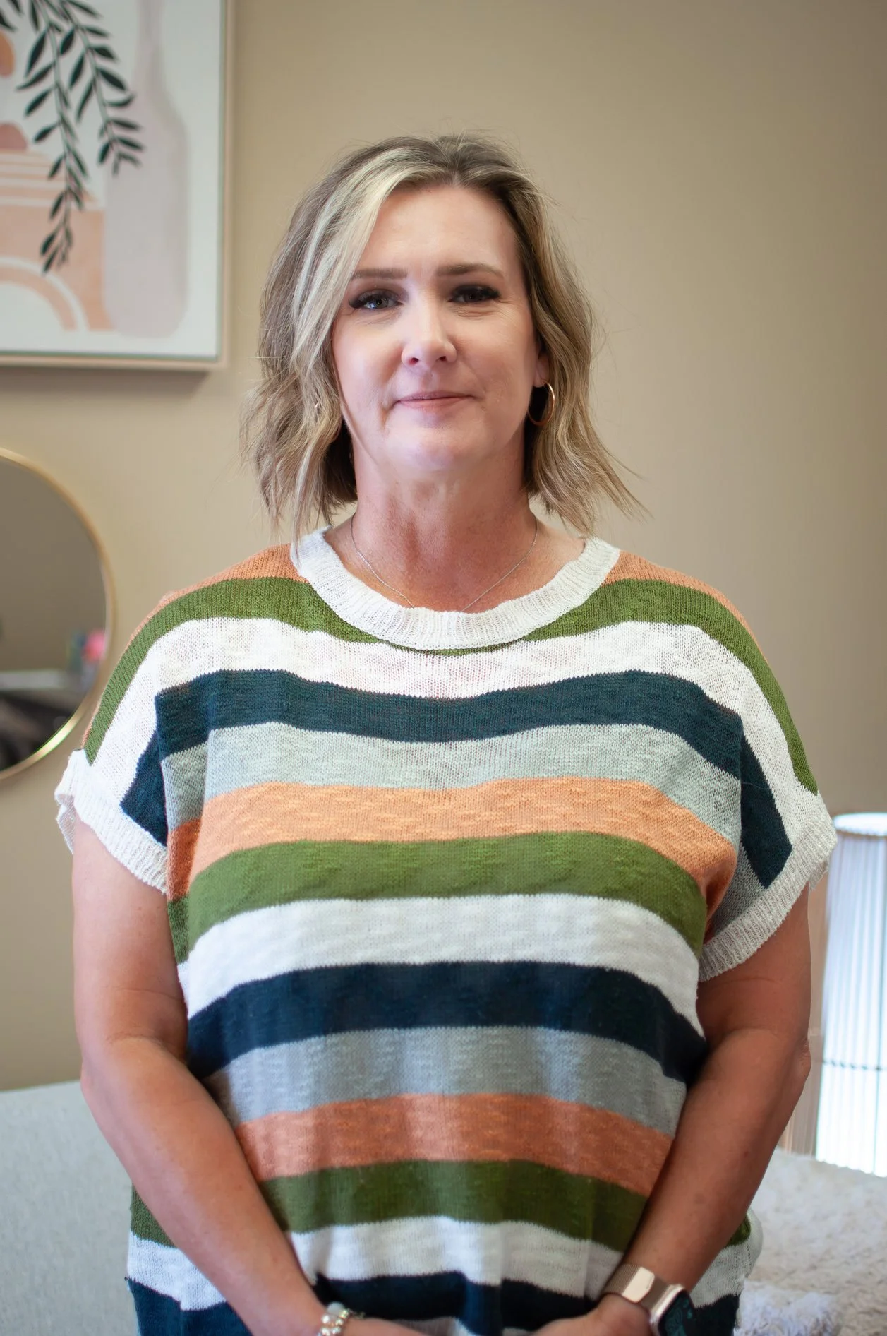 A woman with short blonde hair smiling, wearing a denim shirt and gold hoop earrings, standing in front of two framed wall art pieces featuring abstract leaves.