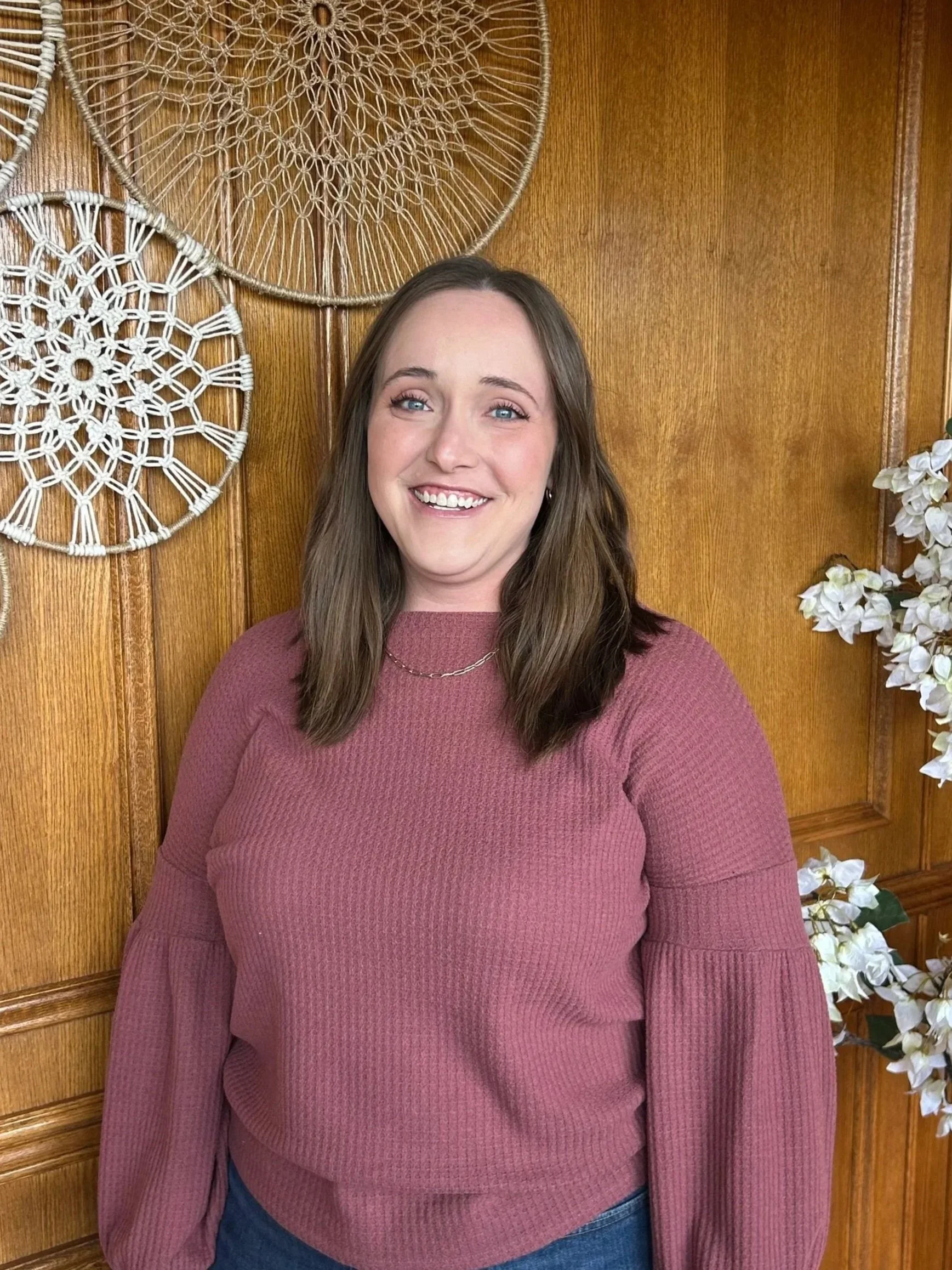 A woman with long brown hair, glasses, and light makeup, wearing a white top and a gold pendant necklace, standing in front of a gray stone wall.