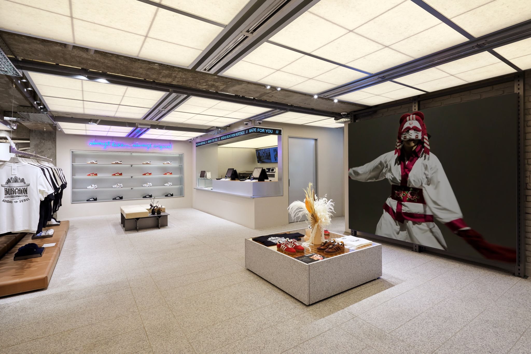 Modern retail store interior with sneakers on display, clothing on racks, a checkout counter, and a large screen showing a traditional dancer.