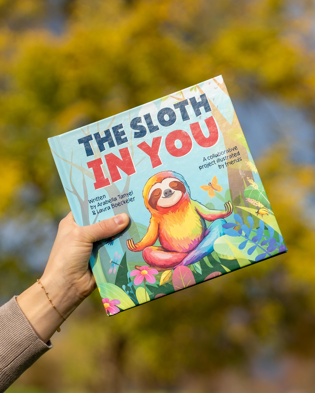 Launch post - The Sloth In You book held in the sky triumphantly by a hand with nature in the background