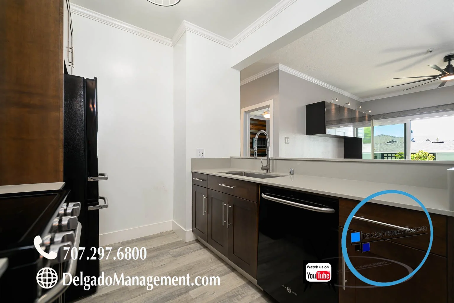 Modern kitchen with dark wooden cabinets, white countertops, stainless steel sink, black appliances, and a view of living area with large window and ceiling fan.