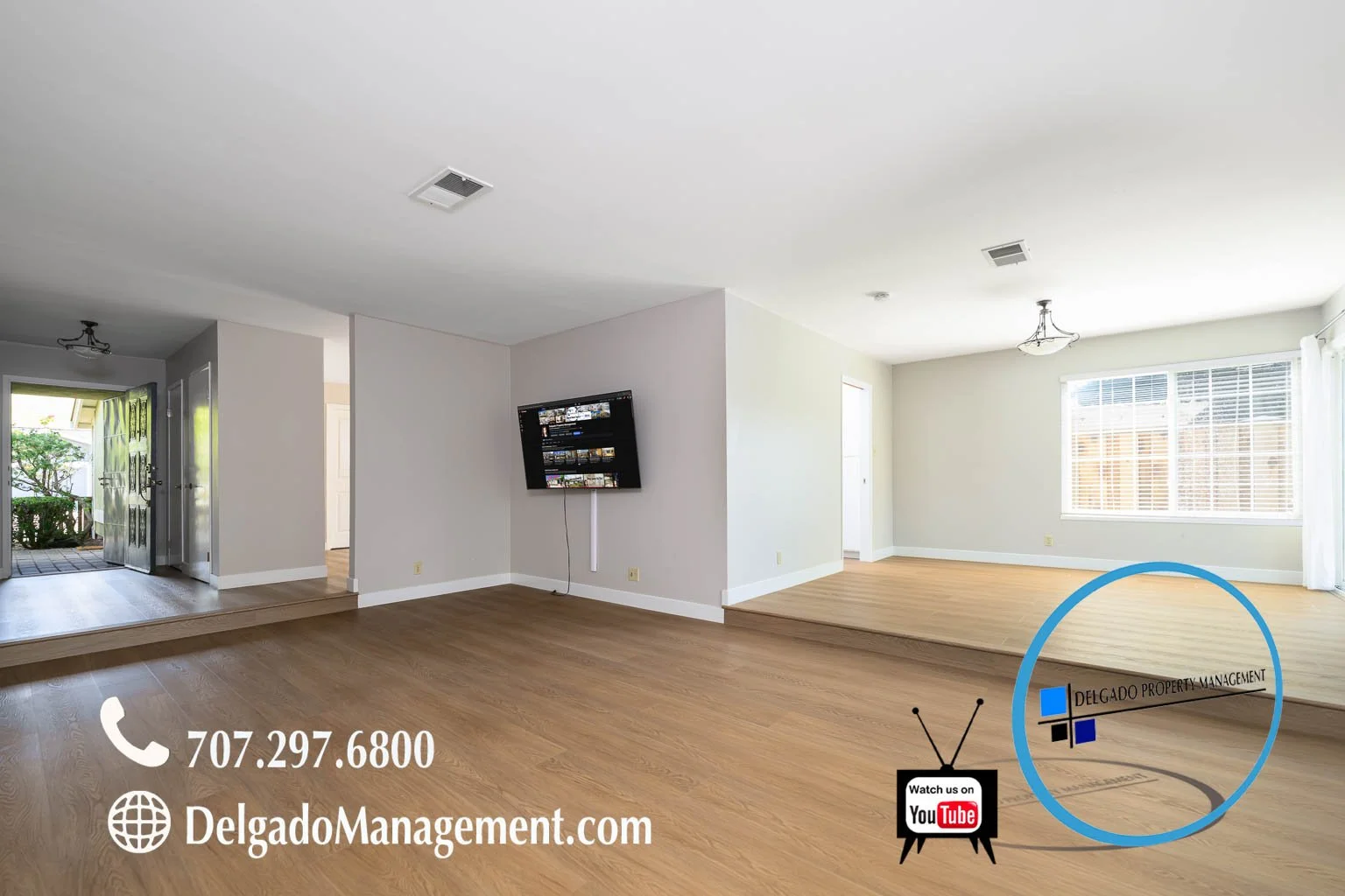 Empty living room with hardwood floors, white walls, large window with blinds, and a TV mounted on a partial wall. Logo and contact information for Delgado Property Management are in the lower right corner.