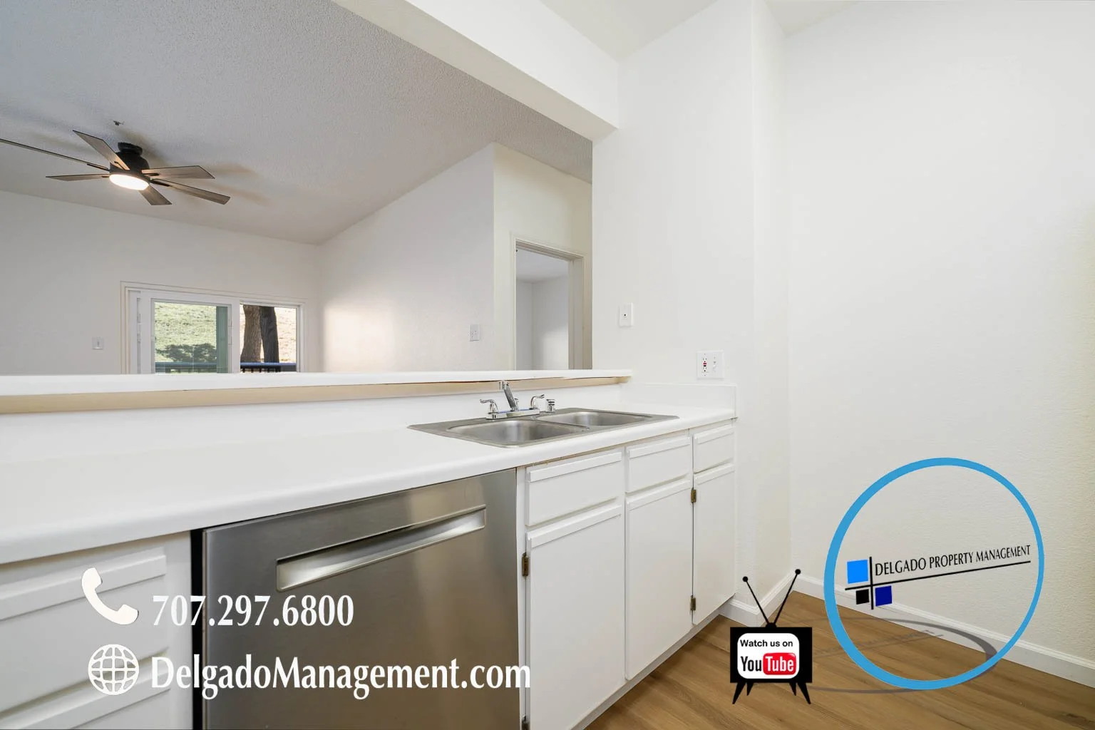 Kitchen with white cabinets and countertop, stainless steel sink, and dishwasher. Background shows a view through a large window to trees and a sliding door. There is a circular graphic with the logo and name 'Delgado Property Management' in the lowe