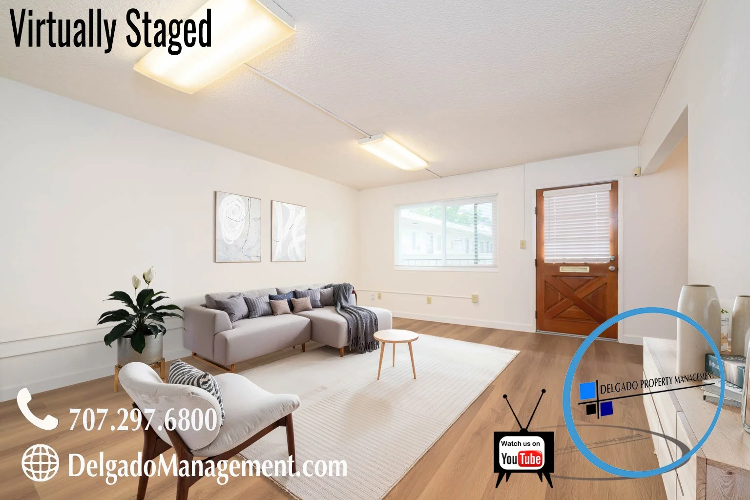 Living room with white walls, wooden flooring, a light gray sectional couch with throw pillows, a white armchair with a black and white pillow, a small wooden coffee table, large window, front door with wooden finish and window blinds, potted plant, 
