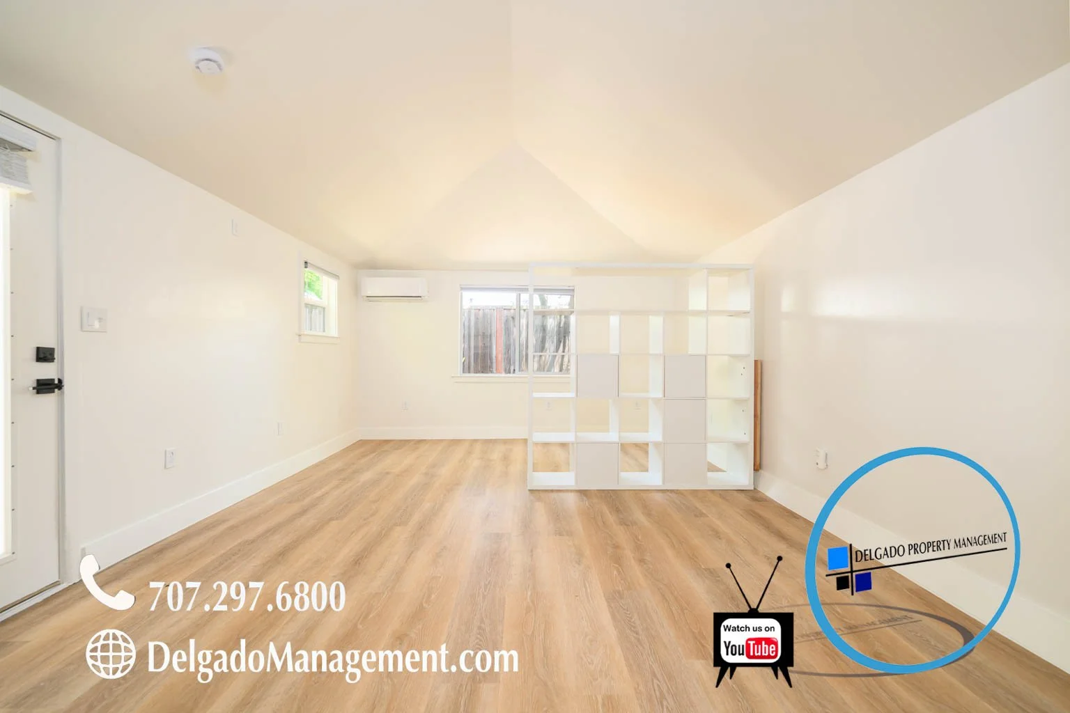 Empty living room with hardwood floors, white walls, a white open shelving unit, and a sliding glass door leading outside, with contact information and branding for Delgado Property Management.