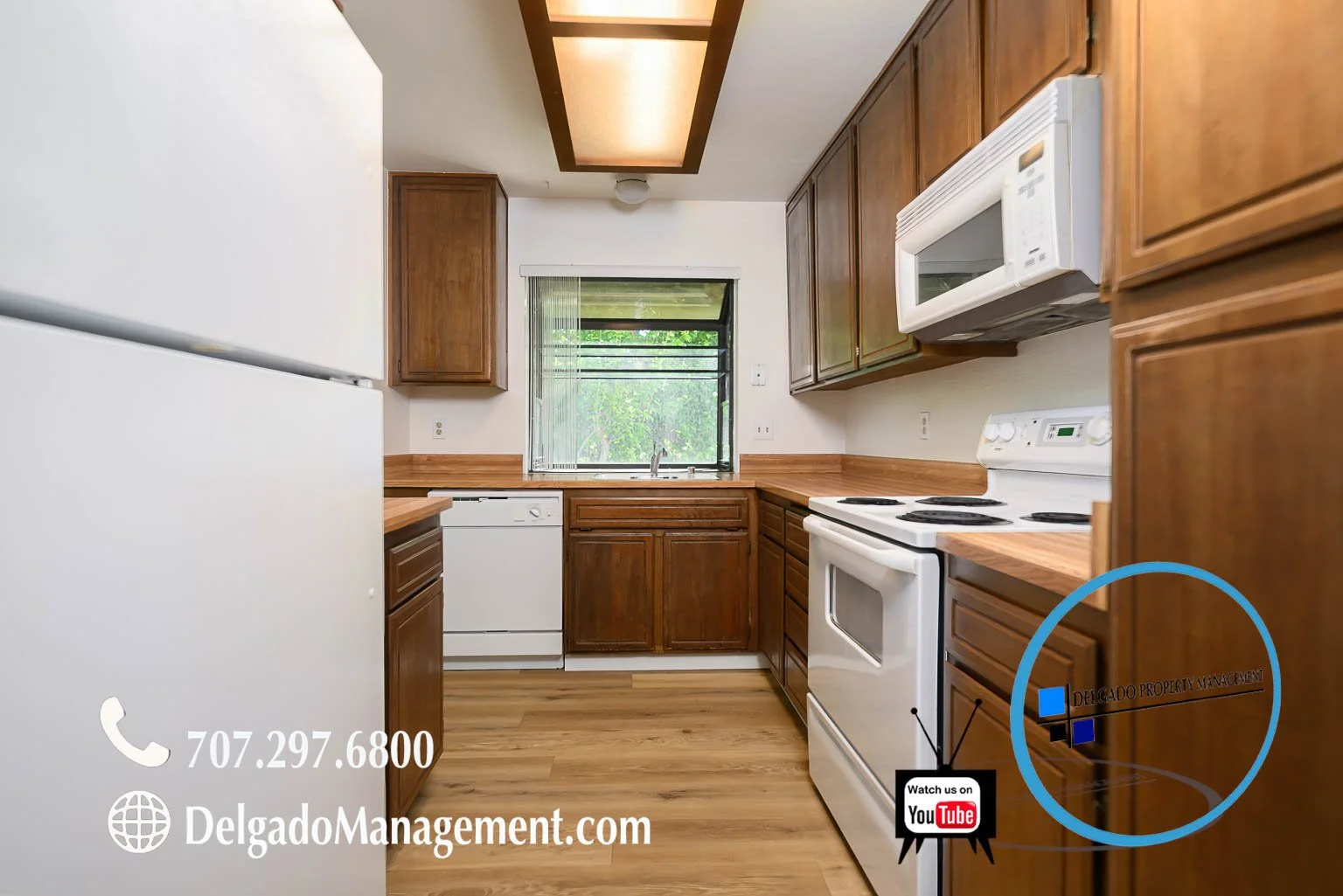 Kitchen with wooden cabinets, white appliances including a refrigerator, dishwasher, microwave, and oven, a window with blinds showing greenery outside, and wood flooring.