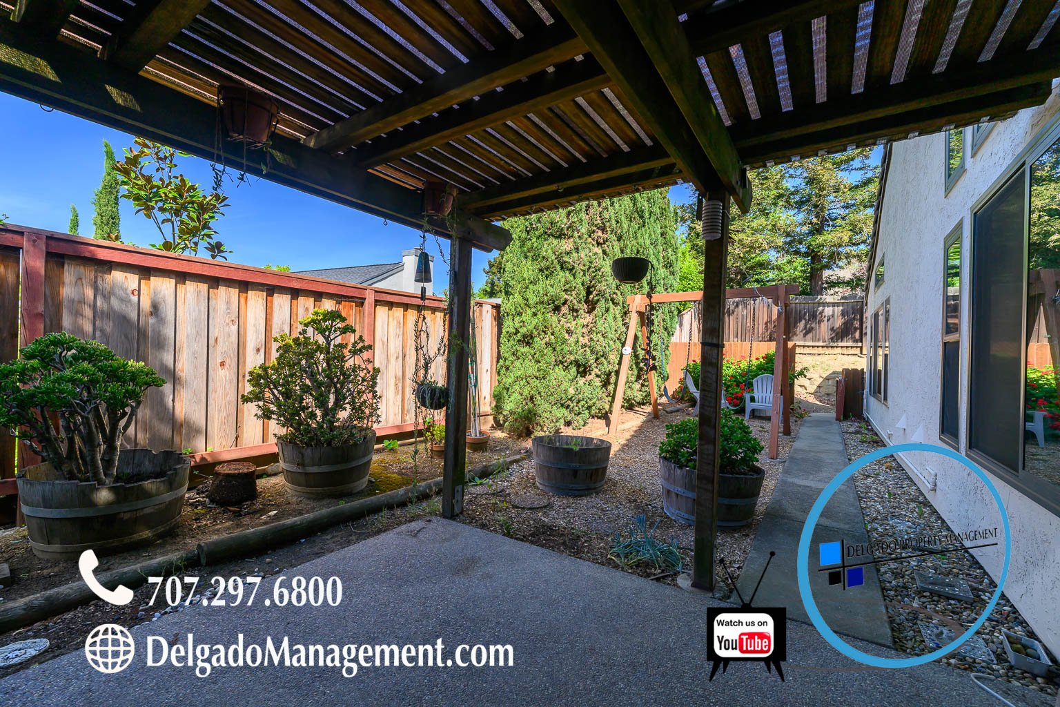 Backyard patio with potted plants, a wooden fence, a house wall on the right, and a swing set in the background.