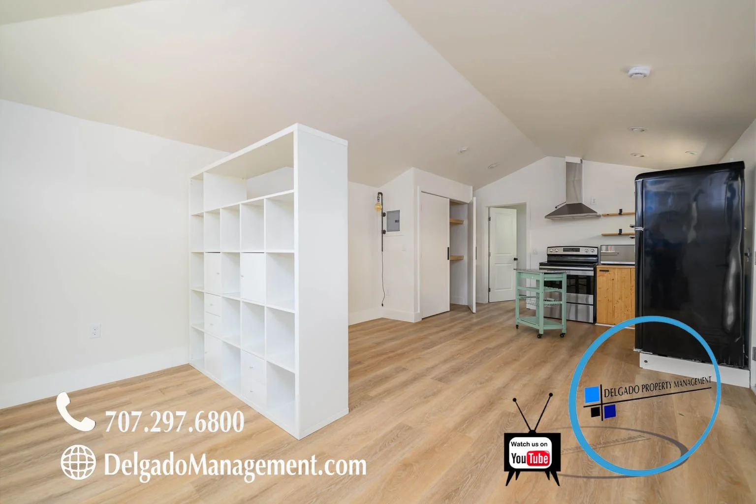 Empty living area with light wood floors, white walls, and a kitchen in the background including a black refrigerator, stove, and wooden shelving. Contact information and website for Delgado Property Management displayed at the bottom, along with You