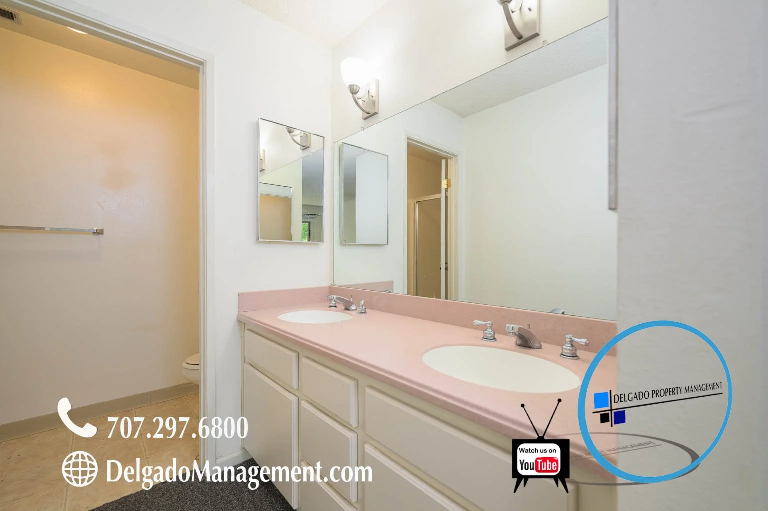 Bathroom with a double sink vanity with pink countertop and two mirrors. There are wall-mounted light fixtures above the mirrors. The bathroom has beige floor tiles, a door to the toilet area, and a Delgado Property Management logo with contact infor