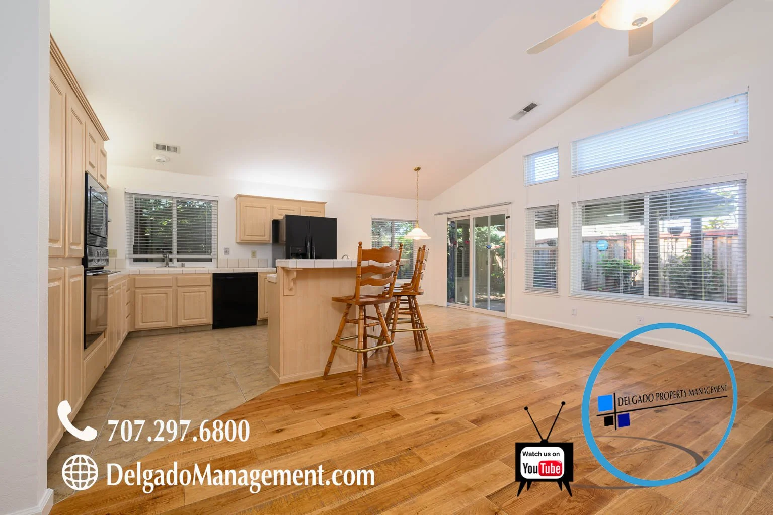 Empty open-concept kitchen and living area with river hardwood floors, beige cabinetry, black appliances, and large windows with blinds, overlooking a fenced backyard.