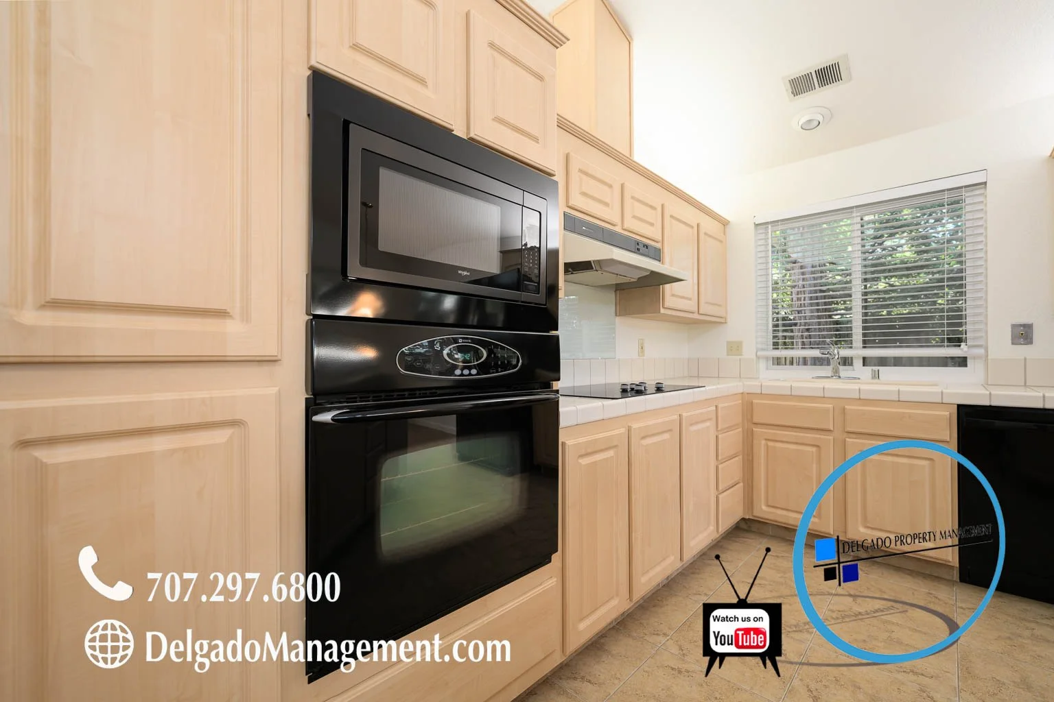 Kitchen with beige cabinets, white tiled countertops, black microwave and oven, window with blinds, and a logo for Delgado Property Management.