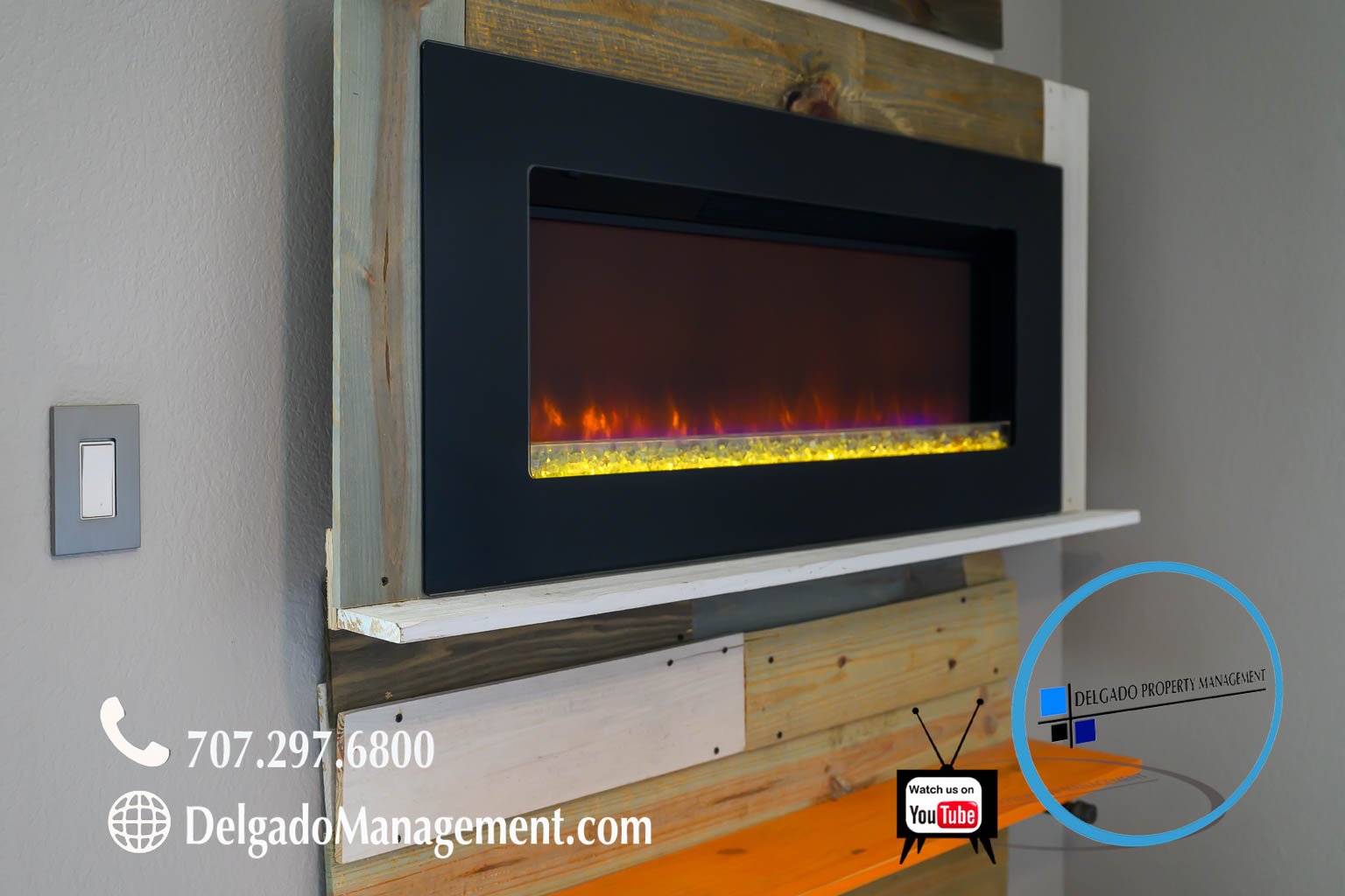 A modern electric fireplace with a black frame, mounted on a piece of wood paneling on a wall. There is a light switch on the wall to the left, and contact information for Delgado Property Management at the bottom left corner, including a phone numbe