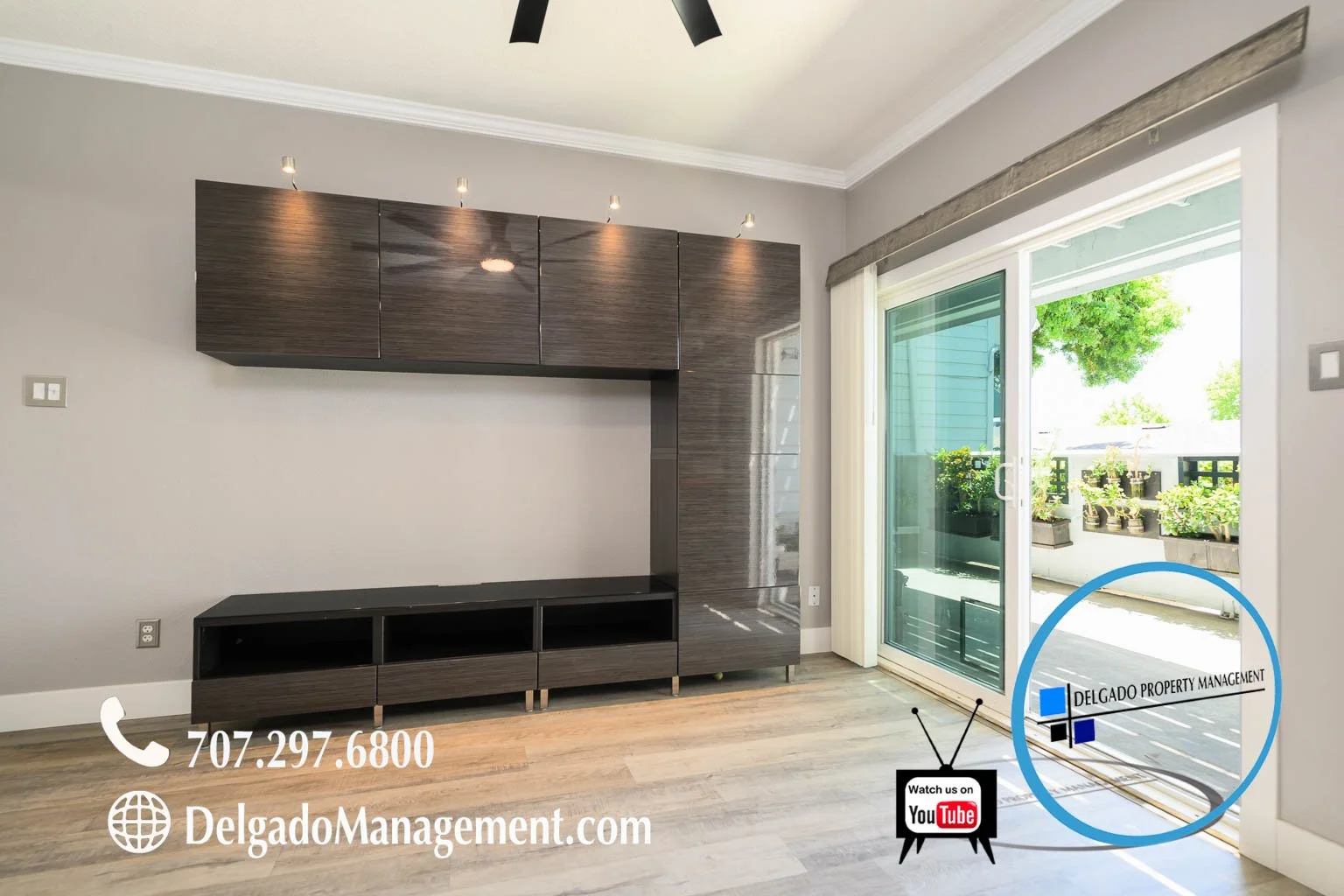 Living room with a wall-mounted dark wood entertainment unit, sliding glass door opening to a balcony with potted plants, and contact information and logo of Delgado Property Management in the lower right corner.