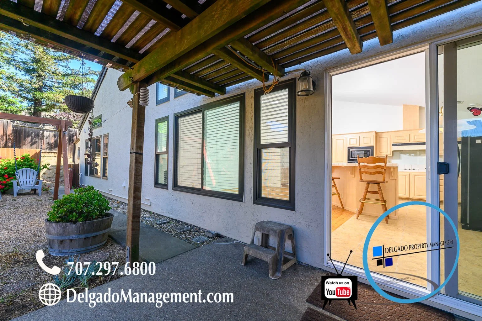 Backyard patio area with a wooden pergola, windowed wall of house, sliding glass door, and a glimpse into a kitchen with wooden cabinets and a chair. There is a potted plant, a small step stool, a swing, and a mailbox on the patio. Contact informatio