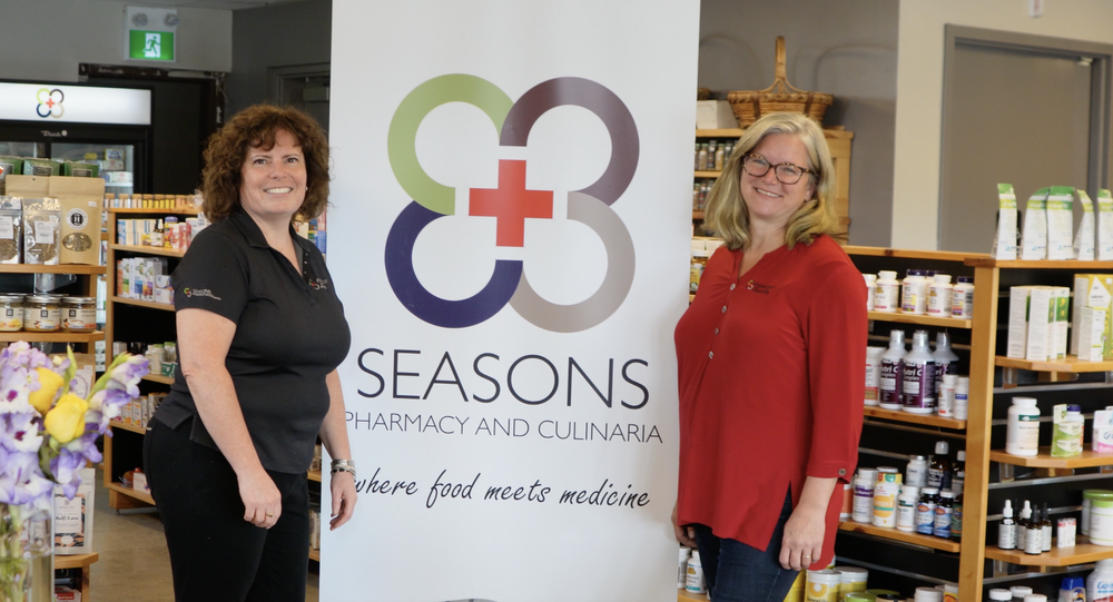 Two pharmacists standing beside a Seasons Pharmacy and Culinaria banner